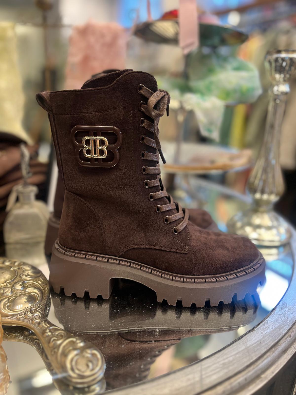 Brun snörkänga i mocka står uppställd på ett runt glassbord och visas i profil, med kraftig räfflad platåsula och metallmärke med bokstäverna “BB” på skaftet; butiksmiljö i mjuk oskärpa.