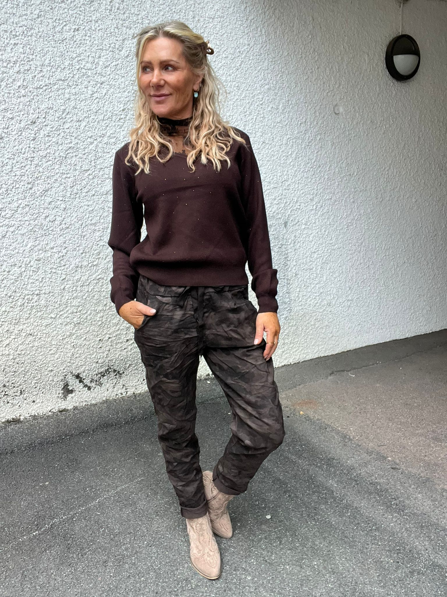 Kvinna med långt blont, vågigt hår står lutad mot en vit, grovputsad vägg och tittar åt sidan. Hon har mörk tröja och kamouflagemönstrade byxor med handen i fickan samt ljusa boots.