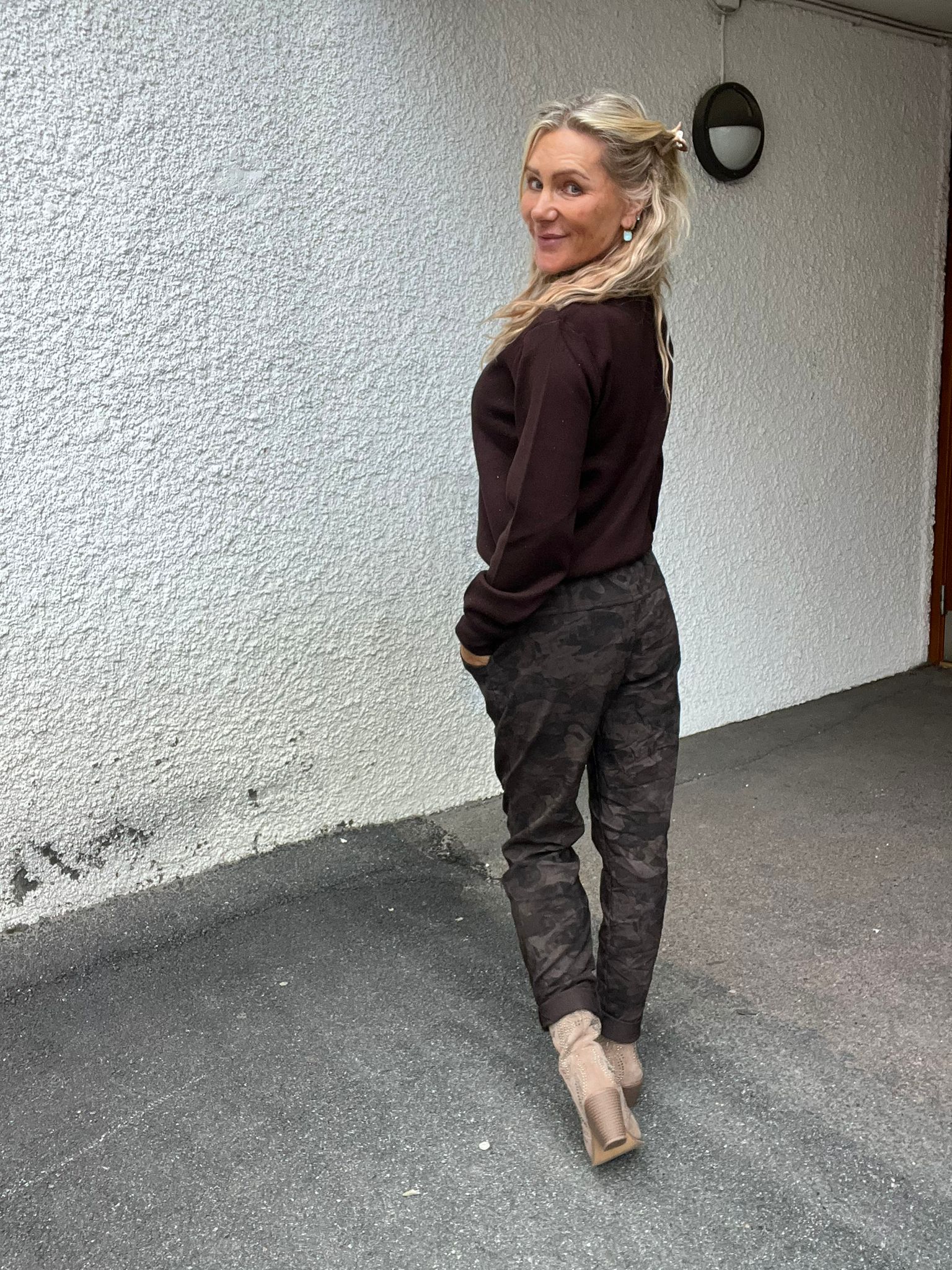 Blond kvinna står på asfalt intill en vit, grovputsad vägg och tittar leende över axeln mot kameran, med händerna i fickorna. Hon bär mörk tröja, kamouflagemönstrade byxor och beige klackskor; vägglampa syns ovanför.
