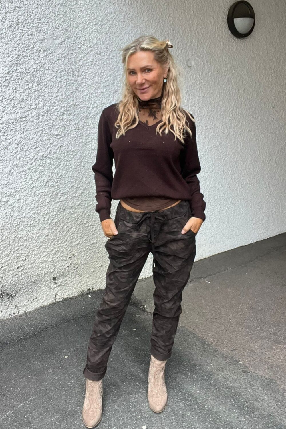 Leende kvinna med långt blont hår står med händerna i fickorna framför en vit putsad vägg utomhus. Hon bär mörk blus med spets vid halsen, kamouflagemönstrade byxor och ljusa boots.