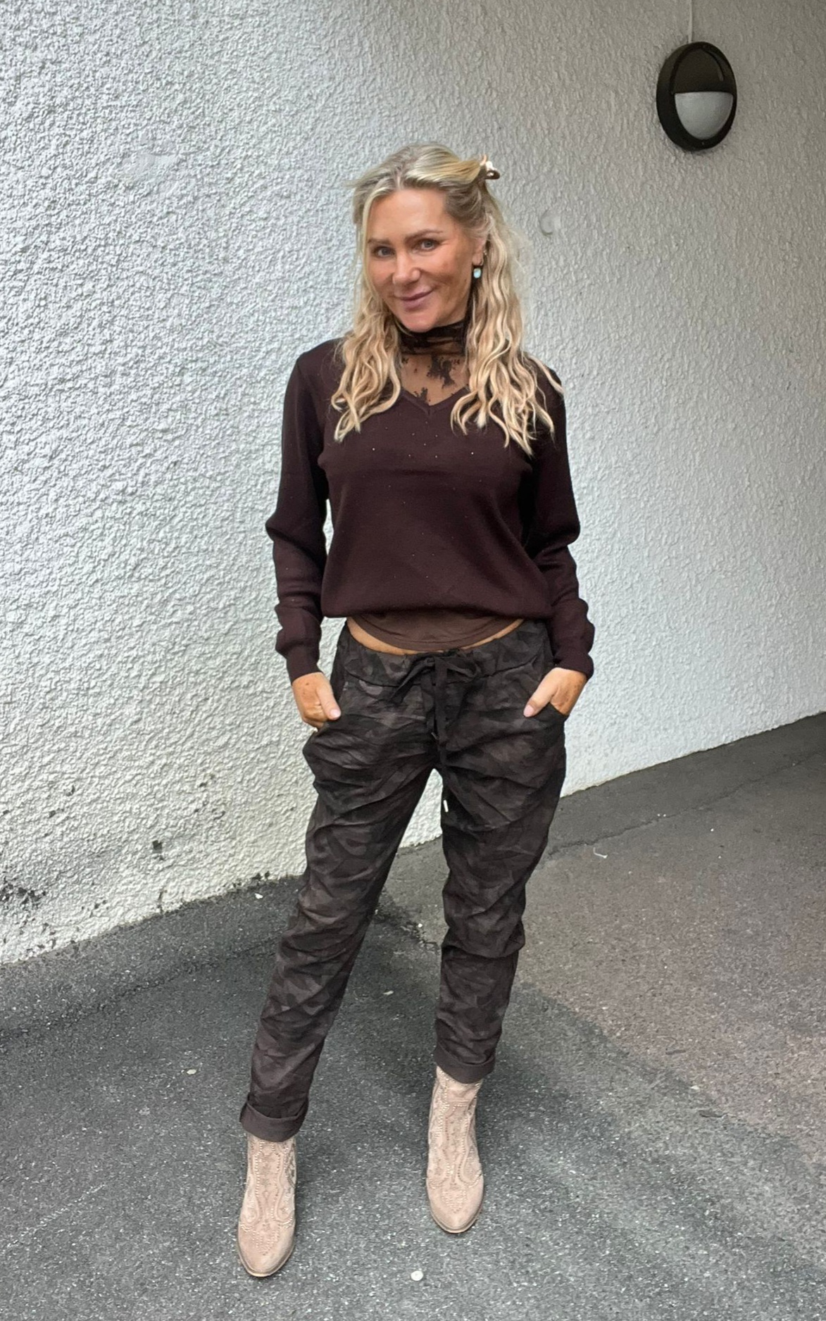 Leende kvinna med långt blont hår står med händerna i fickorna framför en vit putsad vägg utomhus. Hon bär mörk blus med spets vid halsen, kamouflagemönstrade byxor och ljusa boots.