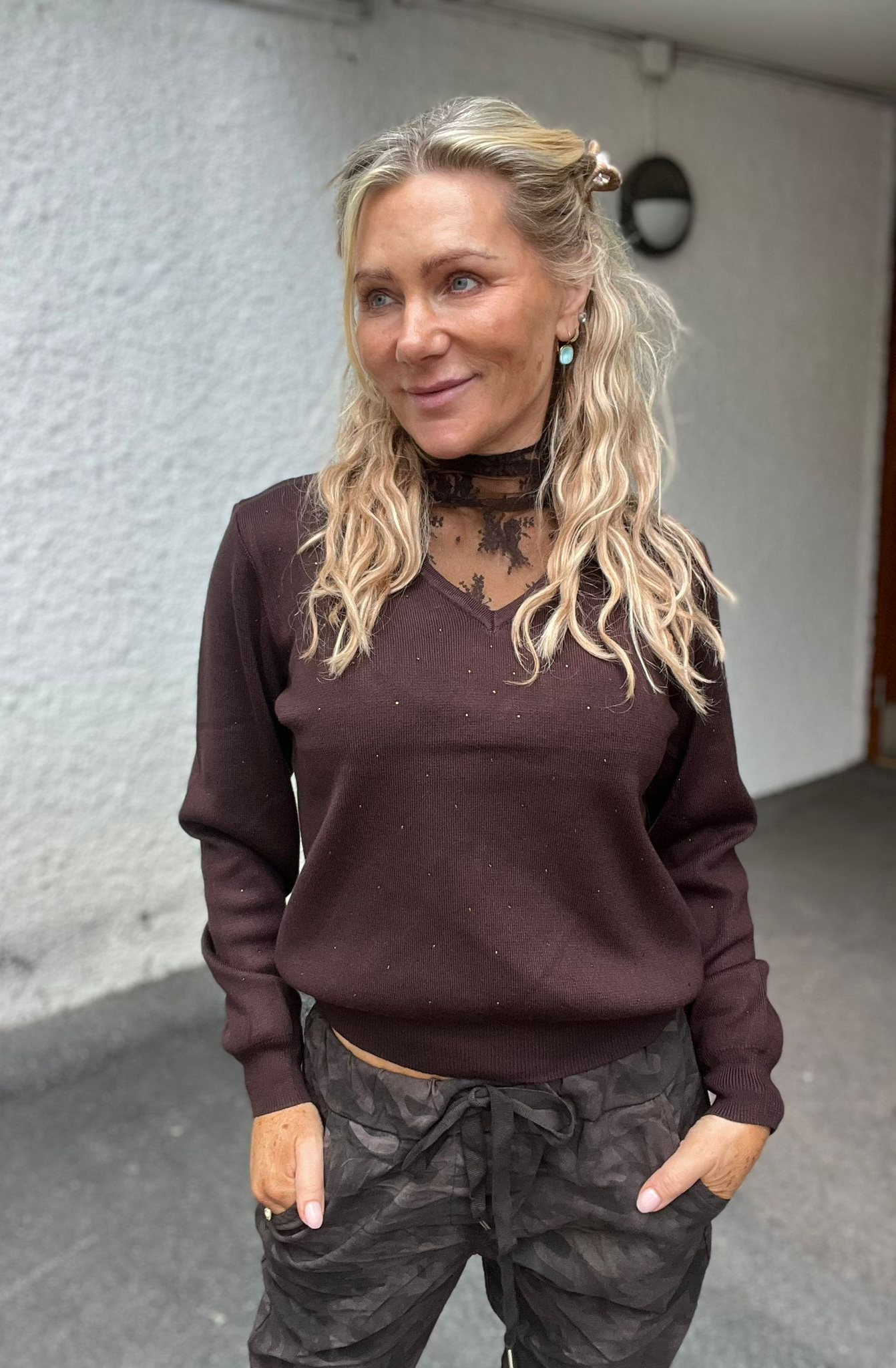 Blond kvinna står avslappnat med händerna i fickorna och ler svagt, blickar åt sidan. Hon bär brun tröja med spetskrage och kamouflagebyxor, mot ljus vägg i en utomhusgång med vägglampa.