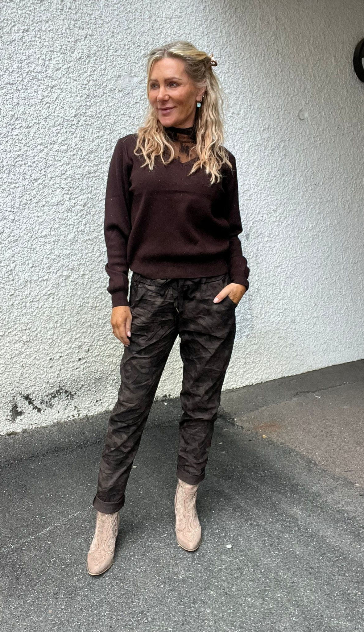 Kvinna med långt blondt hår står avslappnat med ena handen i fickan och ler åt sidan. Hon bär mörk tröja med spetsdetalj vid halsen, mörka mönstrade byxor och ljusa boots mot en vit putsad vägg.