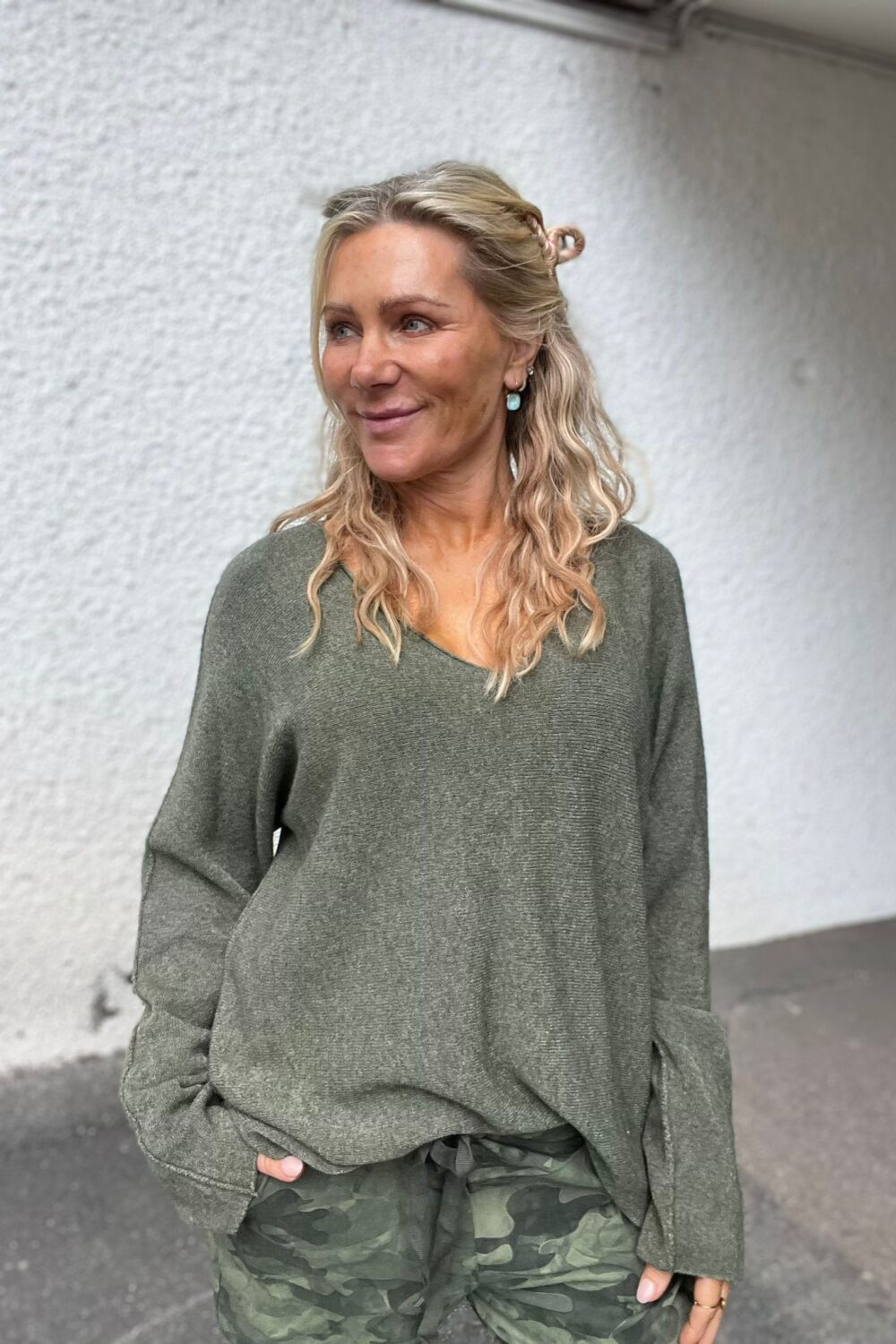Blond kvinna står utomhus mot en ljus, grov vägg och ler medan hon tittar åt sidan. Hon har håret halvt uppsatt, bär grön stickad tröja och kamouflagebyxor med handen i fickan.