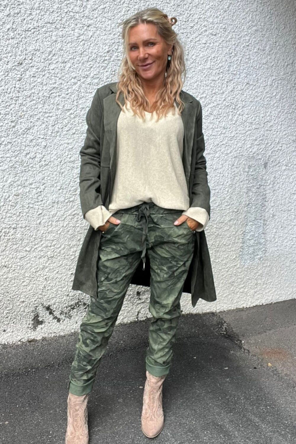 Blond kvinna står lutad mot en vit, grovputsad vägg med händerna i fickorna och ler mot kameran. Hon bär mörkgrön lång kavaj, ljus topp, camouflagemönstrade byxor och ljusa boots på asfalt.