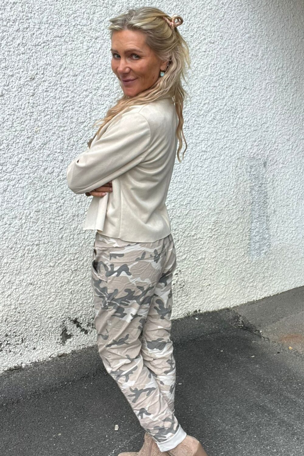 Blond kvinna står i profil med armarna i kors och tittar leende mot kameran. Hon bär ljus kavaj, kamouflagemönstrade byxor och beige boots, framför en vit, grovputsad vägg på asfalt utomhus.