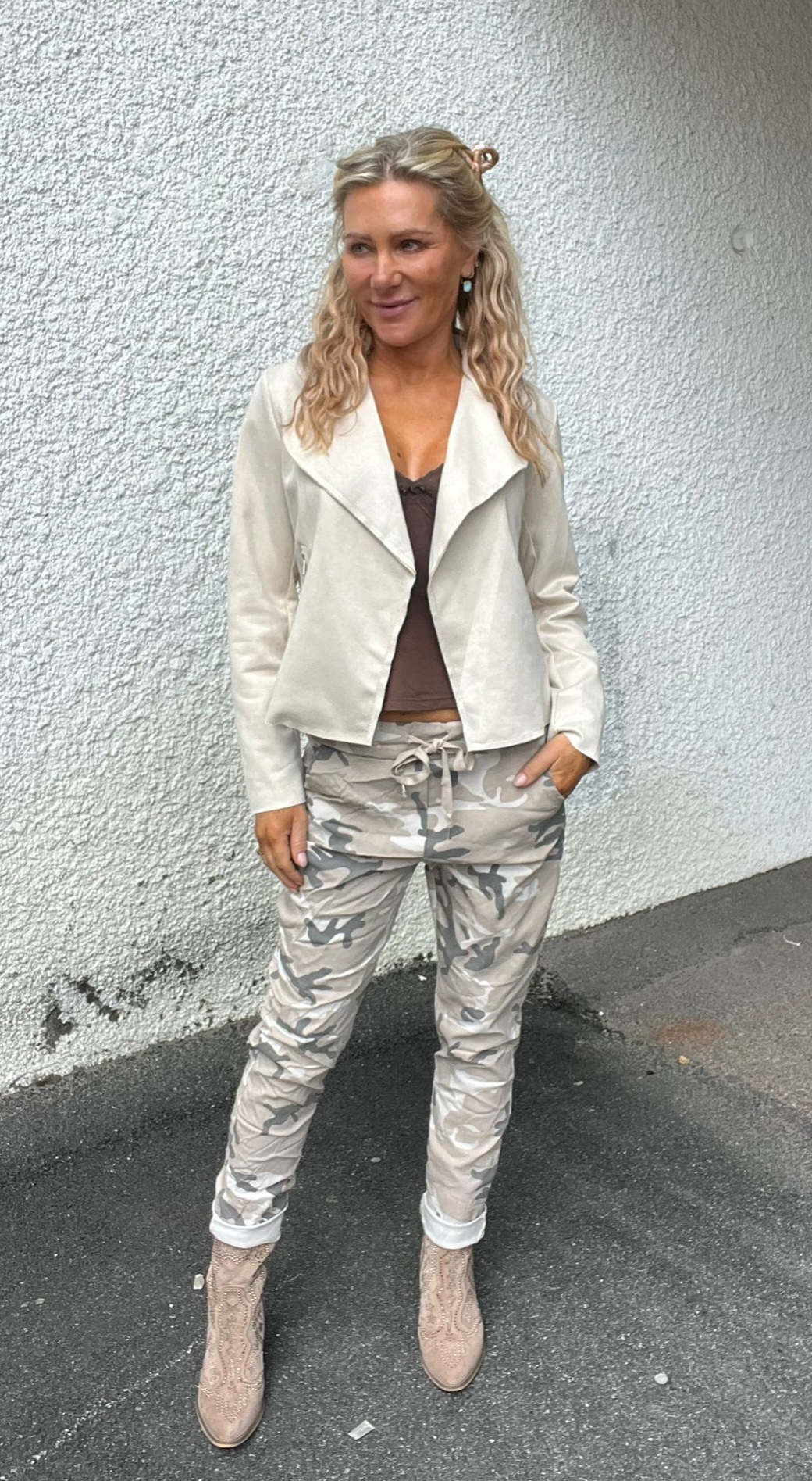 Kvinna står avslappnat med ena handen i fickan framför en vit, grovputsad vägg och tittar åt sidan. Hon bär vit jacka över brun topp, ljusa kamouflagebyxor och beige mönstrade boots.
