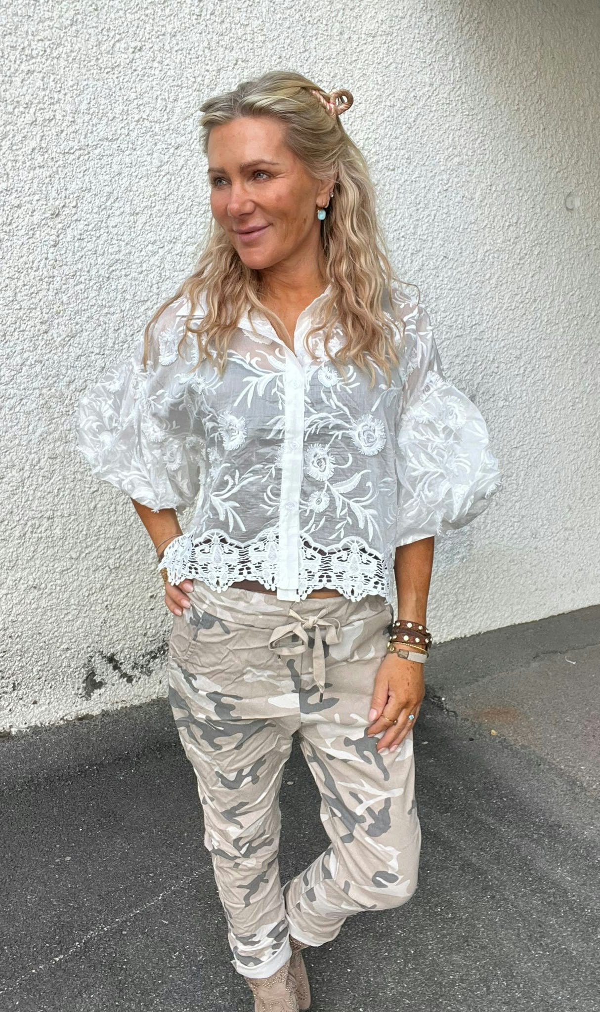 Blond kvinna står lutad mot en vit, grovputsad vägg och ler medan hon tittar åt sidan. Hon bär en vit transparent blus med broderade blommor och kamouflagebyxor, med armband och örhängen.