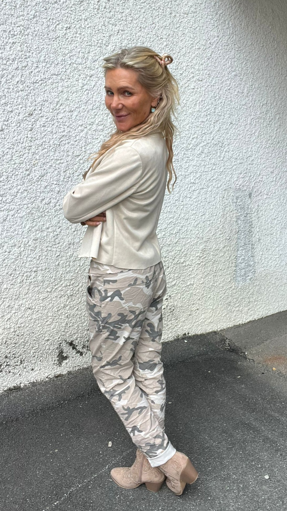 Blond kvinna står i profil med armarna i kors och tittar leende mot kameran. Hon bär ljus kavaj, kamouflagemönstrade byxor och beige boots, framför en vit, grovputsad vägg på asfalt utomhus.