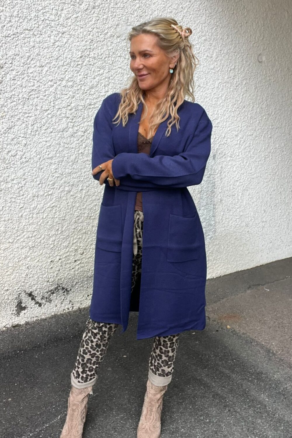 Kvinna står med armarna i kors och ler åt sidan framför en vit, grov vägg utomhus. Hon bär lång blå kappa, leopardmönstrade byxor och ljusa boots, med långt blont hår uppsatt med klämma.