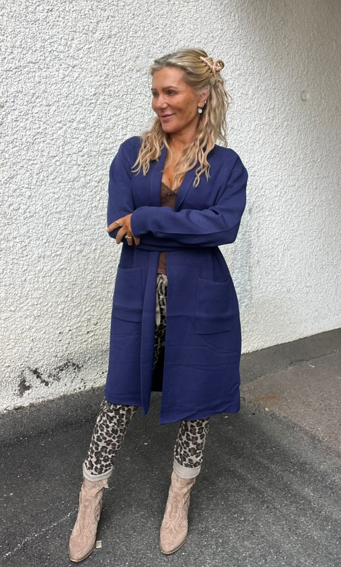 Kvinna står med armarna i kors och ler åt sidan framför en vit, grov vägg utomhus. Hon bär lång blå kappa, leopardmönstrade byxor och ljusa boots, med långt blont hår uppsatt med klämma.