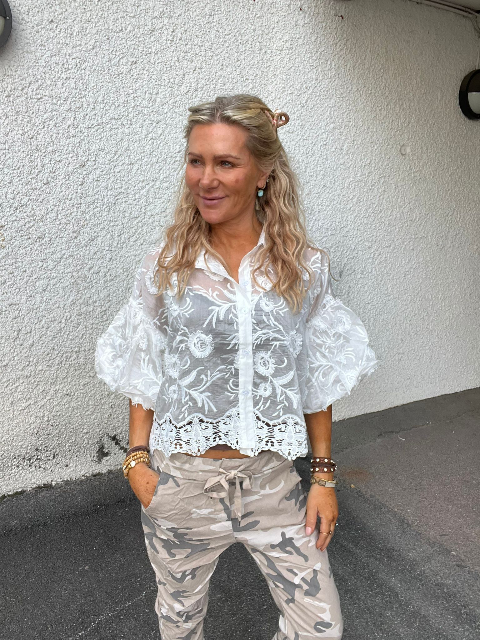 Leende kvinna står utomhus mot en vit, grovputsad vägg och tittar åt sidan. Hon bär vit, genomskinlig broderad blus med volangärmar och ljusa kamouflagebyxor, med armband och örhängen.