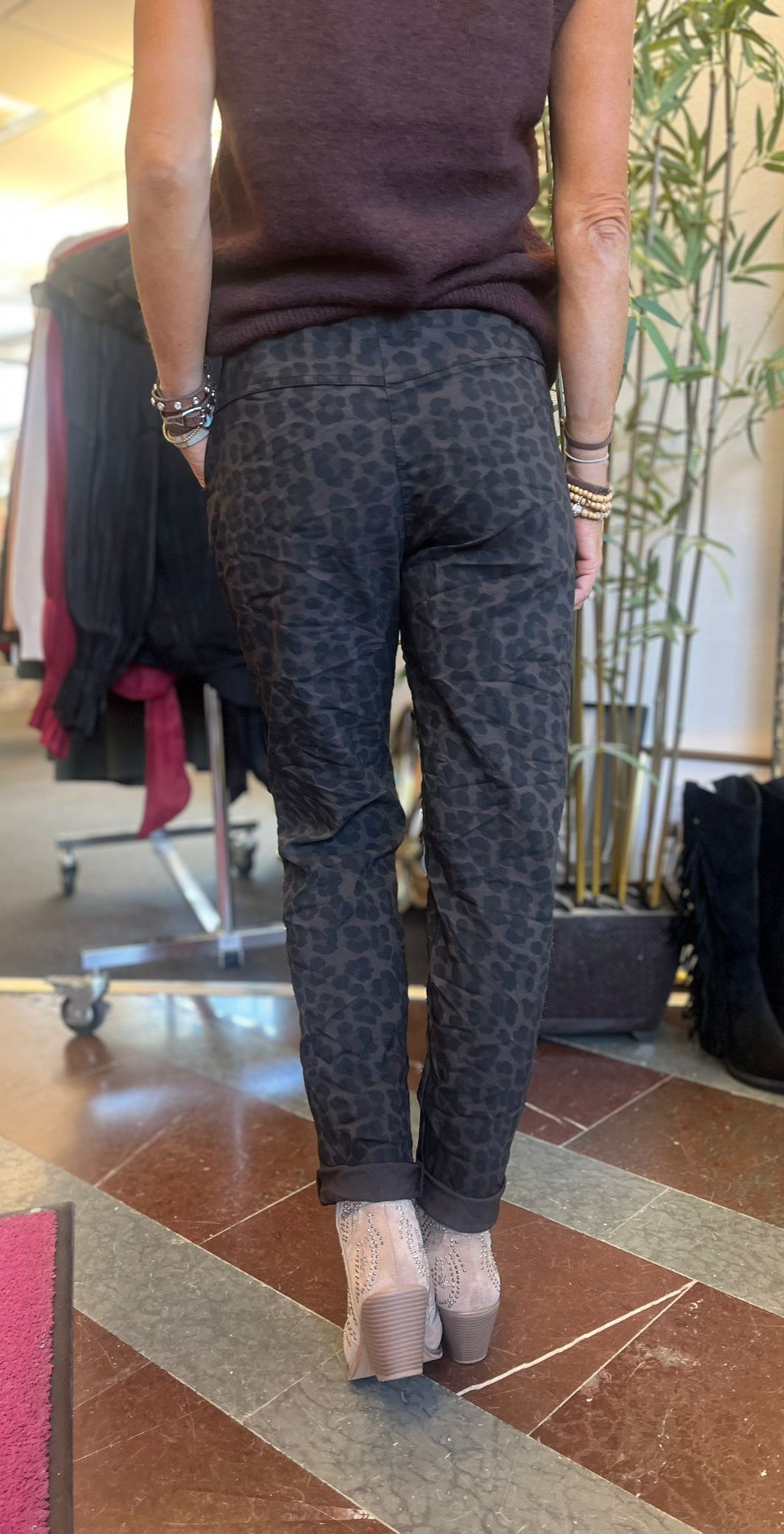 Person sedd bakifrån står inomhus och visar en outfit med mörka leopardmönstrade byxor, svart topp och ljusa ankelboots med klack. Armband på handlederna; i bakgrunden syns klädställning och en bambuväxt.