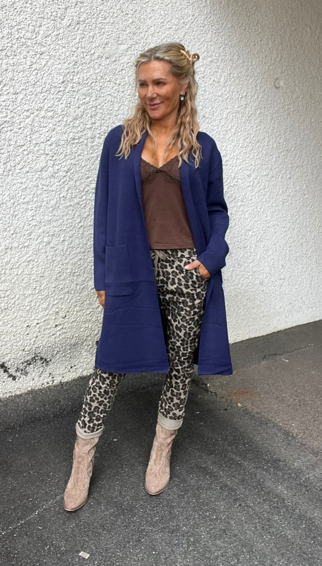 Blond kvinna står avslappnat mot en vit, grovputsad vägg och tittar åt sidan. Hon bär lång blå kofta, brun topp, leopardmönstrade byxor med uppvik och ljusa cowboyboots; ena handen i fickan.