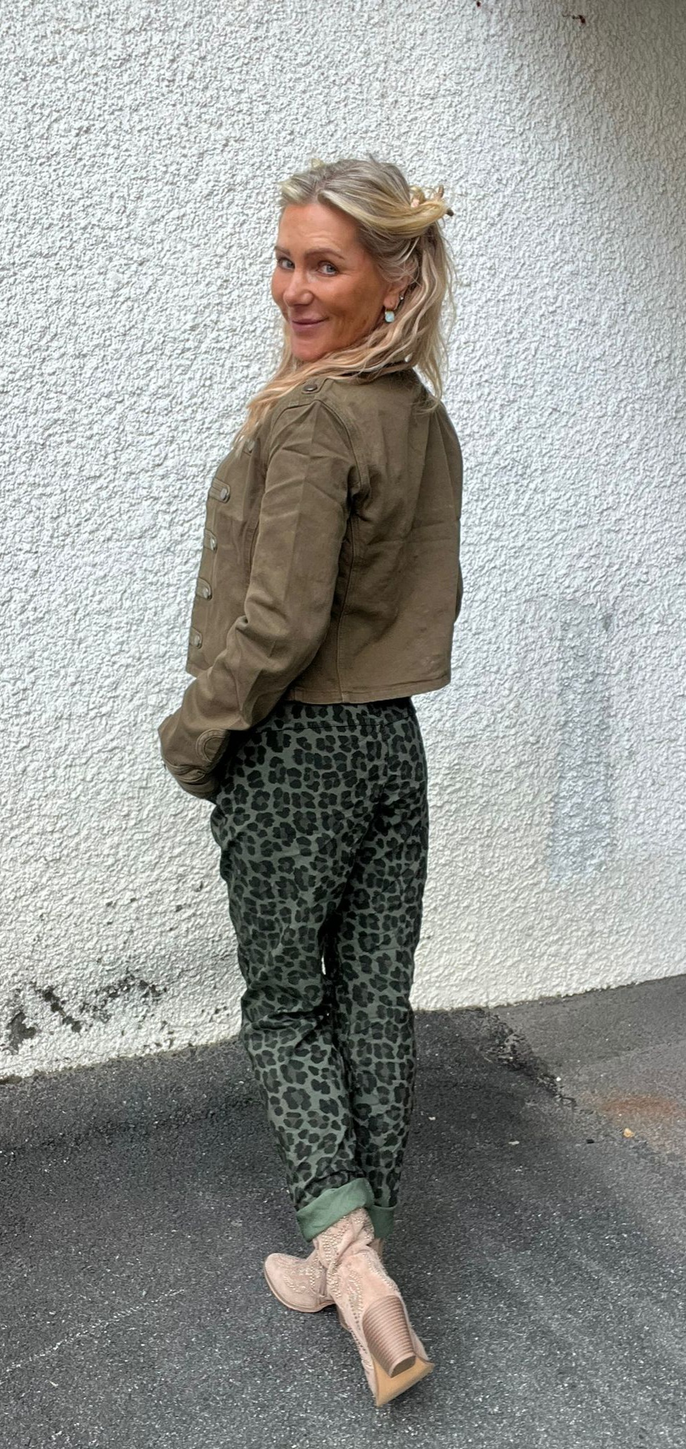 Kvinna med blont halvt uppsatt hår står i profil och tittar leende över axeln mot kameran. Hon bär olivgrön jacka, leopardmönstrade byxor och beige boots, framför en vit grovputsad vägg utomhus.