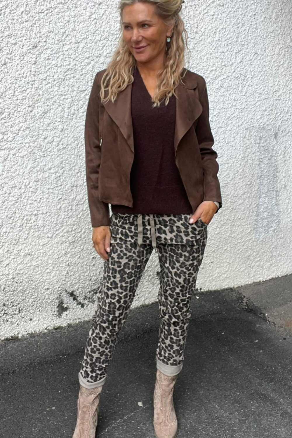 Kvinna står avslappnat mot en vit, grovputsad vägg och tittar åt sidan, med ena handen i fickan. Hon bär brun jacka och topp, leopardmönstrade byxor med snörning samt beige mönstrade boots utomhus.