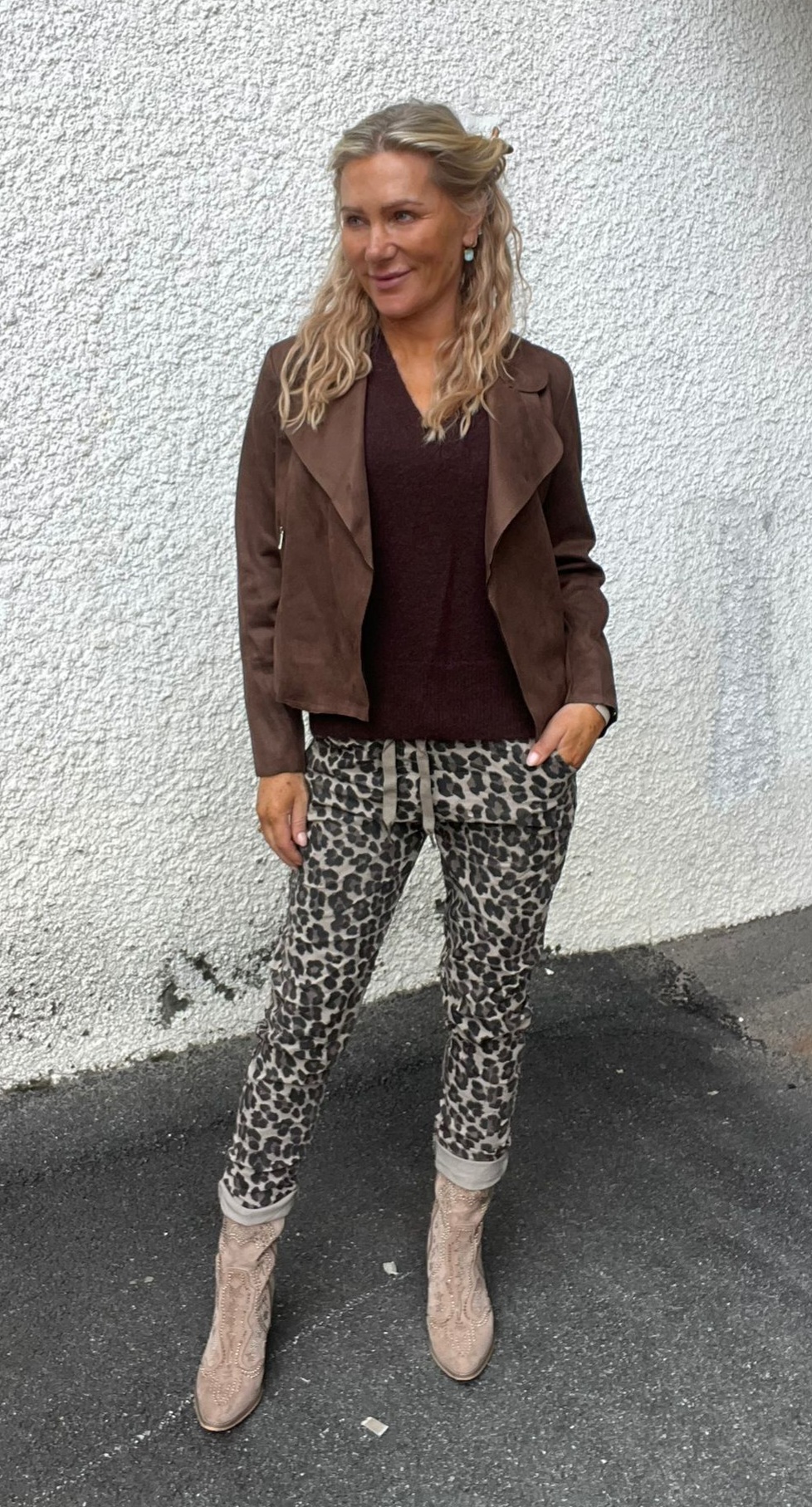 Kvinna med vågigt blont hår står utomhus mot en vit putsad vägg och ser åt sidan med ett leende. Hon bär brun jacka och tröja, leopardmönstrade byxor och beige boots på asfalt.