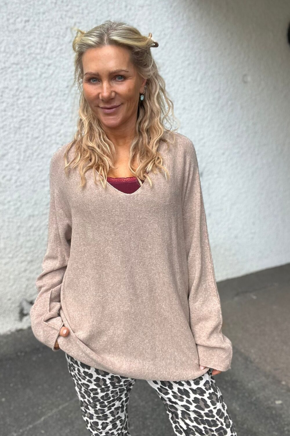 Leende kvinna med långt blont, vågigt hår står avslappnat med händerna i fickorna. Hon bär beige stickad tröja över vinröd topp och leopardmönstrade byxor, framför en ljus, grovputsad vägg utomhus.
