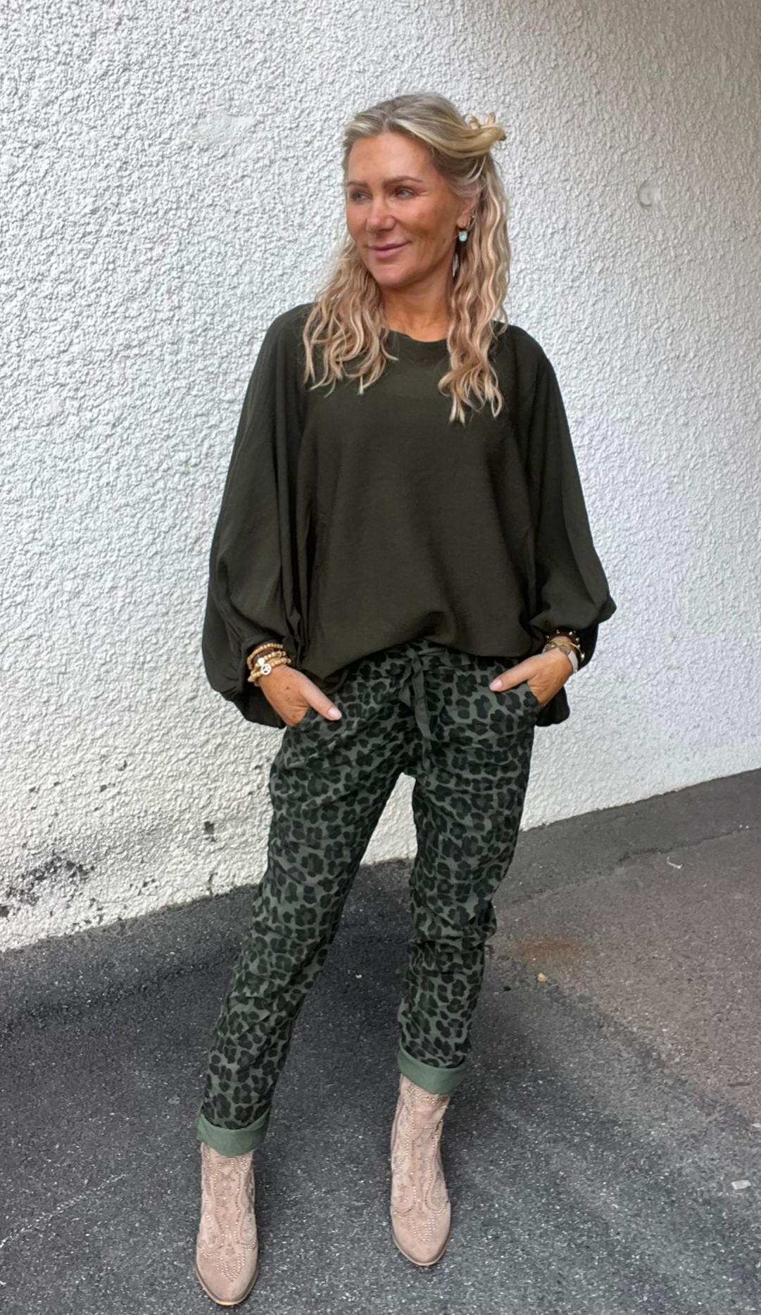 Kvinna med blont, vågigt hår står avslappnat med händerna i fickorna och ler lätt. Hon bär mörkgrön, vidärmad blus, leopardmönstrade byxor och ljusa cowboyboots, mot en vit, grovputsad vägg utomhus.