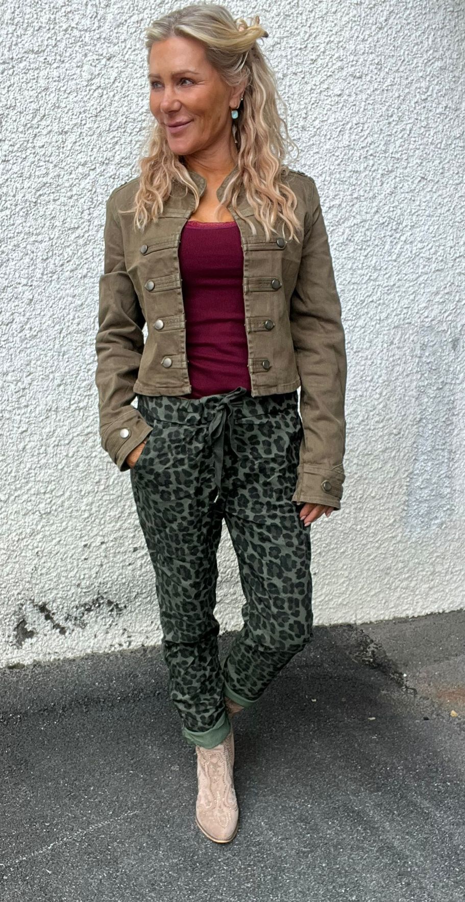 Kvinna står avslappnat med ena handen i fickan och tittar åt sidan, mot en vit putsad vägg. Hon bär kort olivgrön jacka, vinröd topp, leopardmönstrade byxor med knytband och ljusa boots.