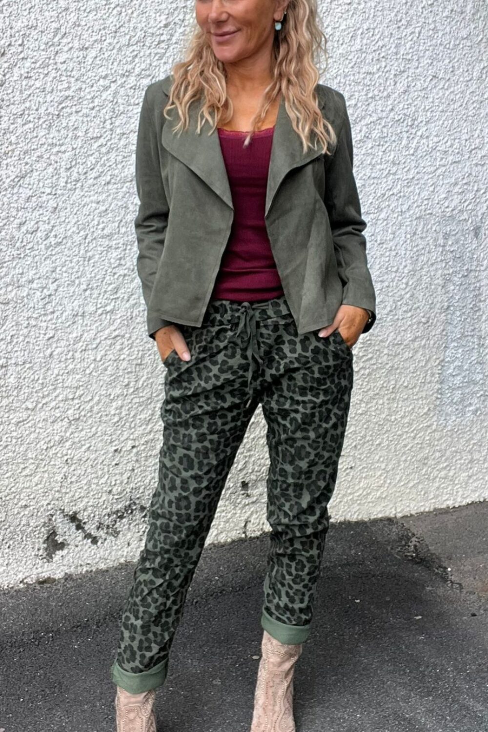 Kvinna med blont, vågigt hår står avslappnat med händerna i fickorna och ler åt sidan. Hon bär olivgrön jacka, vinröd topp, leopardmönstrade byxor och ljusa cowboyboots mot en vit putsad vägg.