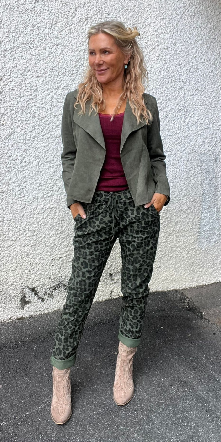 Kvinna med blont, vågigt hår står avslappnat med händerna i fickorna och ler åt sidan. Hon bär olivgrön jacka, vinröd topp, leopardmönstrade byxor och ljusa cowboyboots mot en vit putsad vägg.