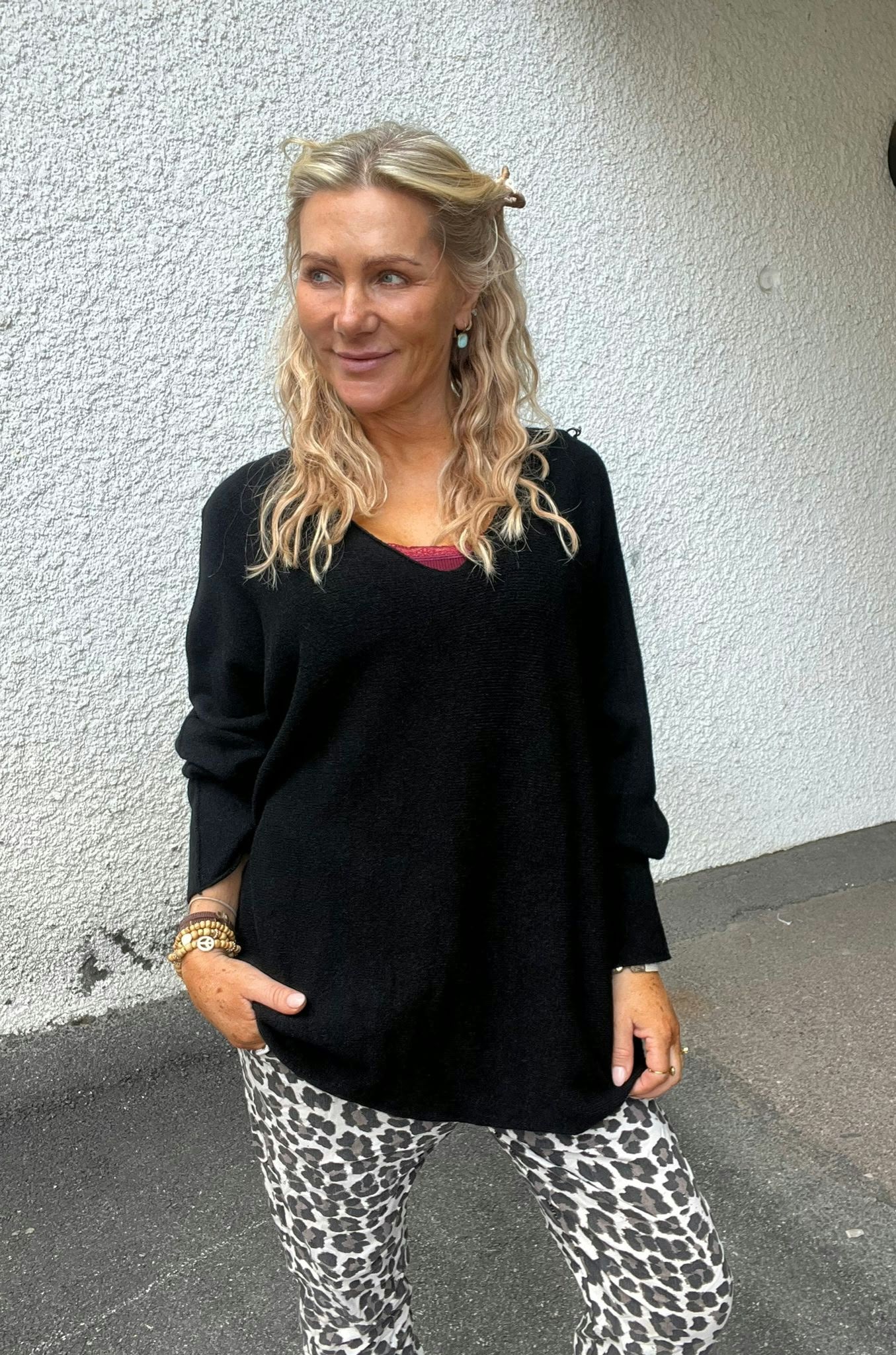 Blond kvinna står utomhus mot en vit, grovputsad vägg och ler medan hon tittar åt sidan. Hon bär svart långärmad tröja med rött linne som syns, leopardmönstrade byxor och armband.
