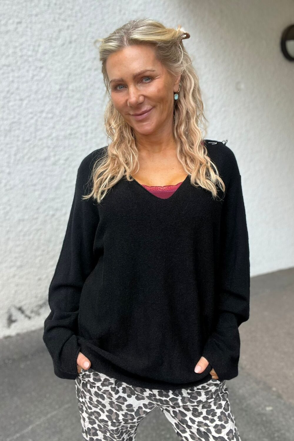 Blond kvinna med vågigt hår ler mot kameran och håller händerna i fickorna. Hon bär svart tröja med röd spetskant och leopardmönstrade byxor, stående utomhus framför en ljus vägg.