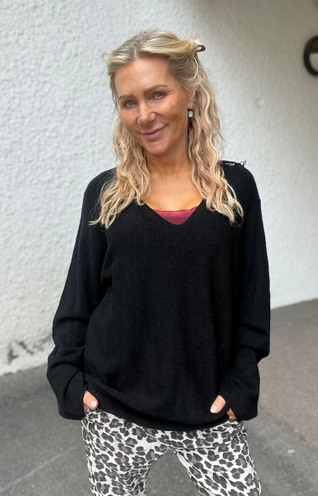 Blond kvinna med vågigt hår ler mot kameran och håller händerna i fickorna. Hon bär svart tröja med röd spetskant och leopardmönstrade byxor, stående utomhus framför en ljus vägg.
