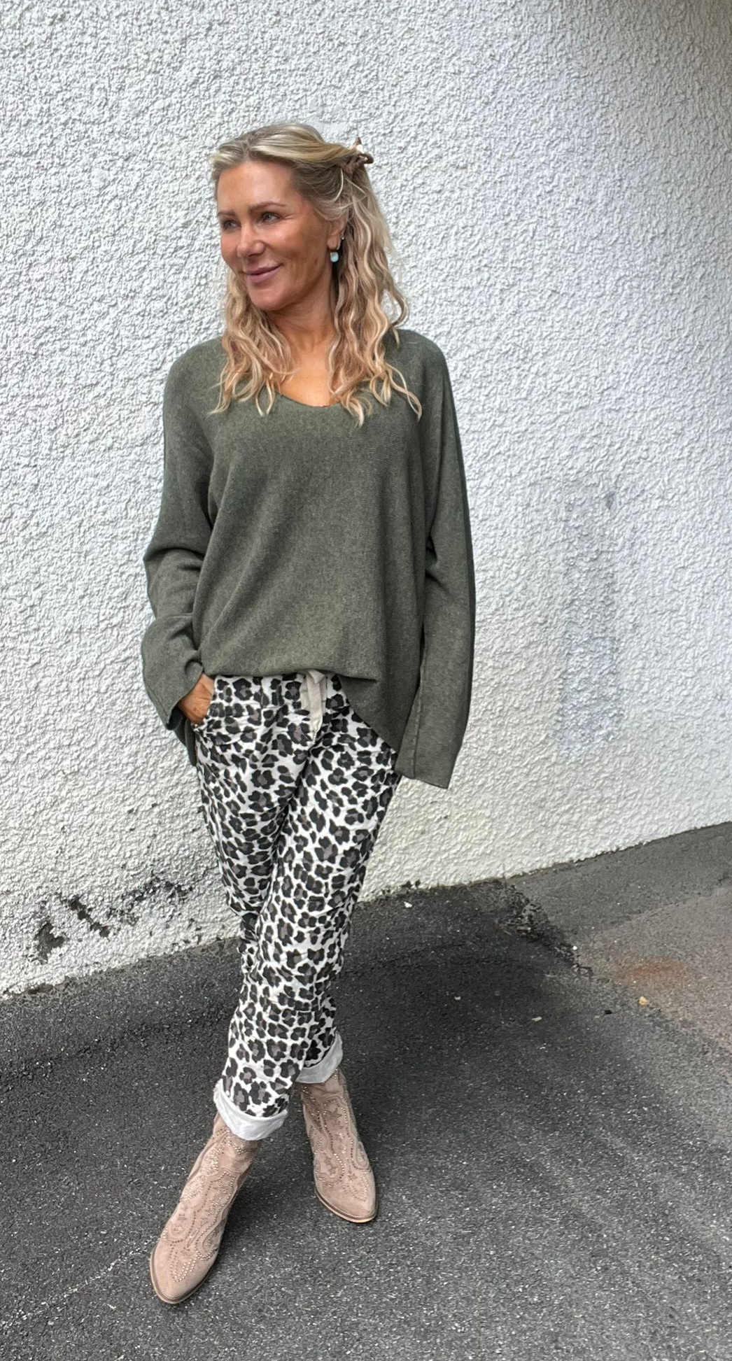 Kvinna med långt blont hår står avslappnat mot en vit, grovputsad vägg och ler åt sidan. Hon har olivgrön stickad tröja, leopardmönstrade byxor och ljusbeige boots; ena handen i fickan.