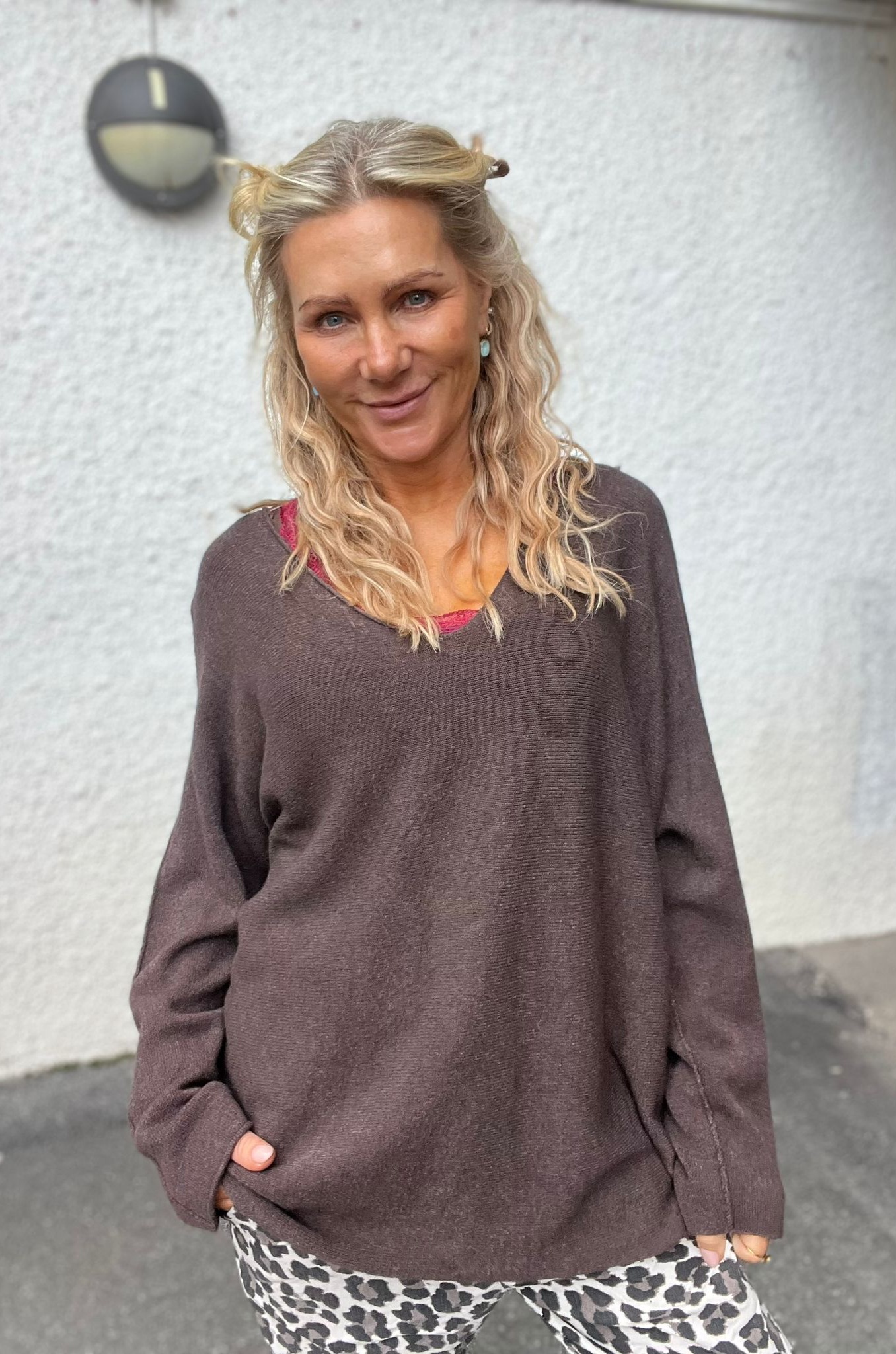 Blond kvinna med vågigt hår ler mot kameran och står med händerna i fickorna. Hon bär brun stickad tröja över en rosa topp och mönstrade byxor, framför en ljus vägg med rund utelampa.