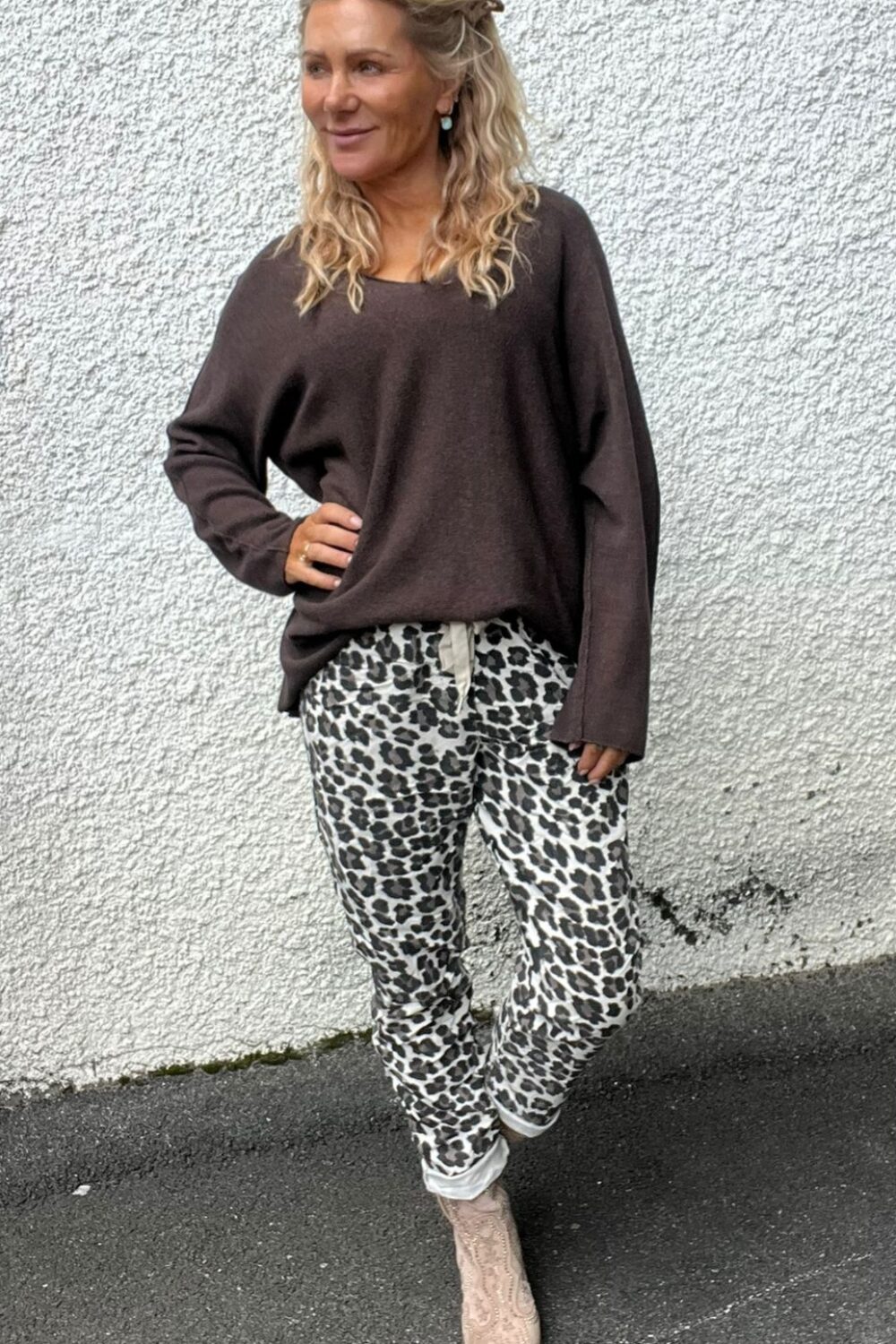 Kvinna med långt, lockigt blont hår står med handen på höften och tittar åt sidan, klädd i mörk oversized tröja och leopardmönstrade byxor med snörning samt ljusa boots, framför vit putsad vägg utomhus.