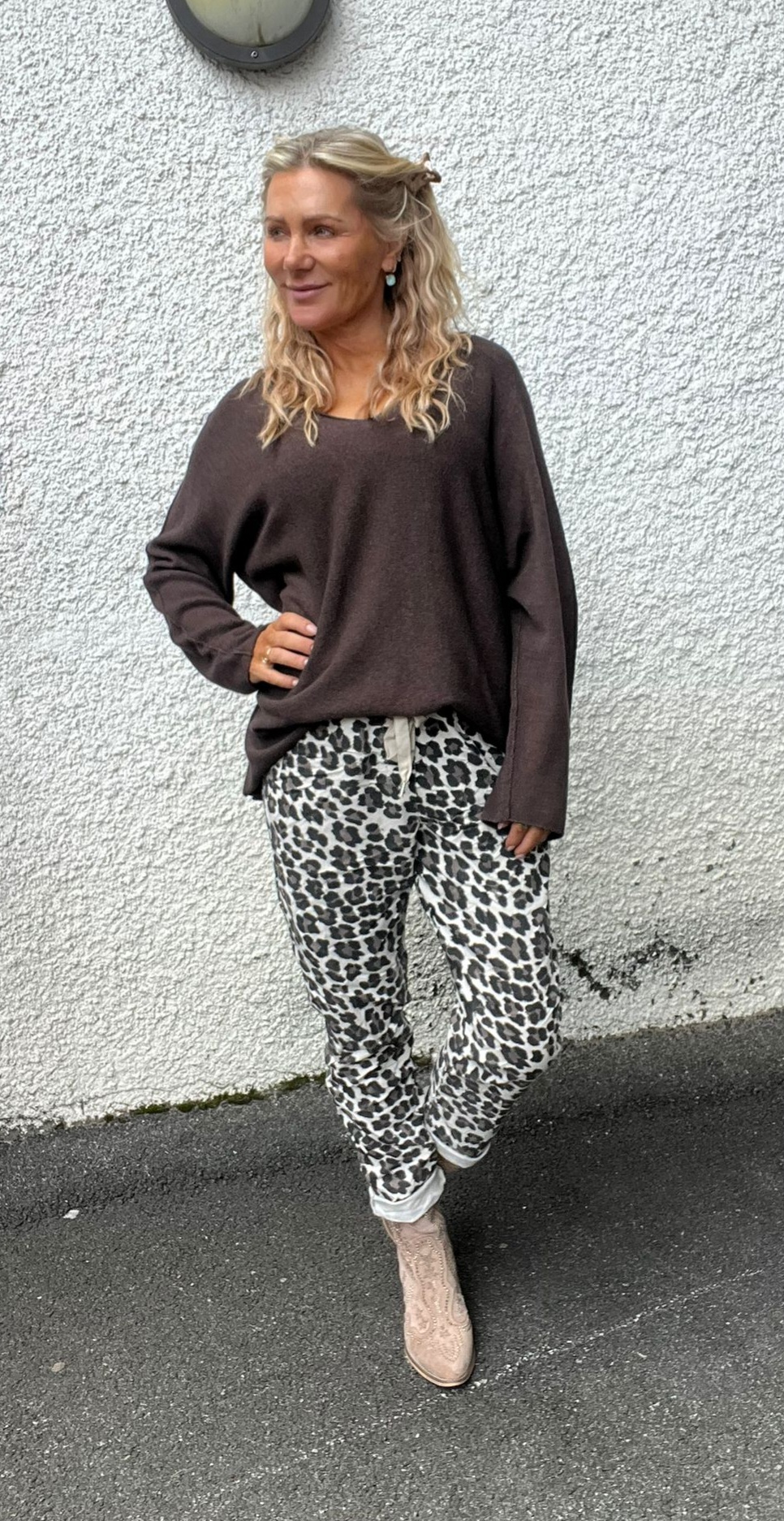 Blond kvinna står avslappnat med ena handen på höften och tittar åt sidan, klädd i brun oversize-tröja och leopardmönstrade byxor med snörning. Hon står mot en vit grovputsad vägg på asfalt, med runda lampan ovanför.