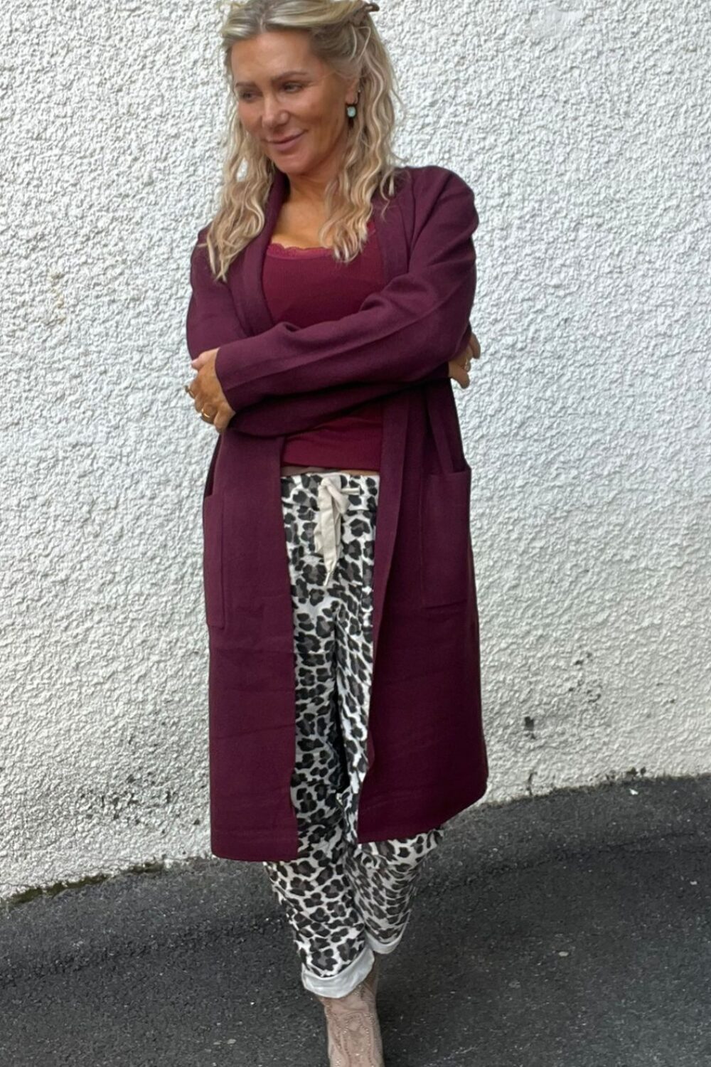 Kvinna står med armarna i kors och lutar sig lätt mot en vit, grovputsad vägg utomhus. Hon bär vinröd lång kofta och linne, leopardmönstrade byxor samt beige boots, och ler åt sidan.