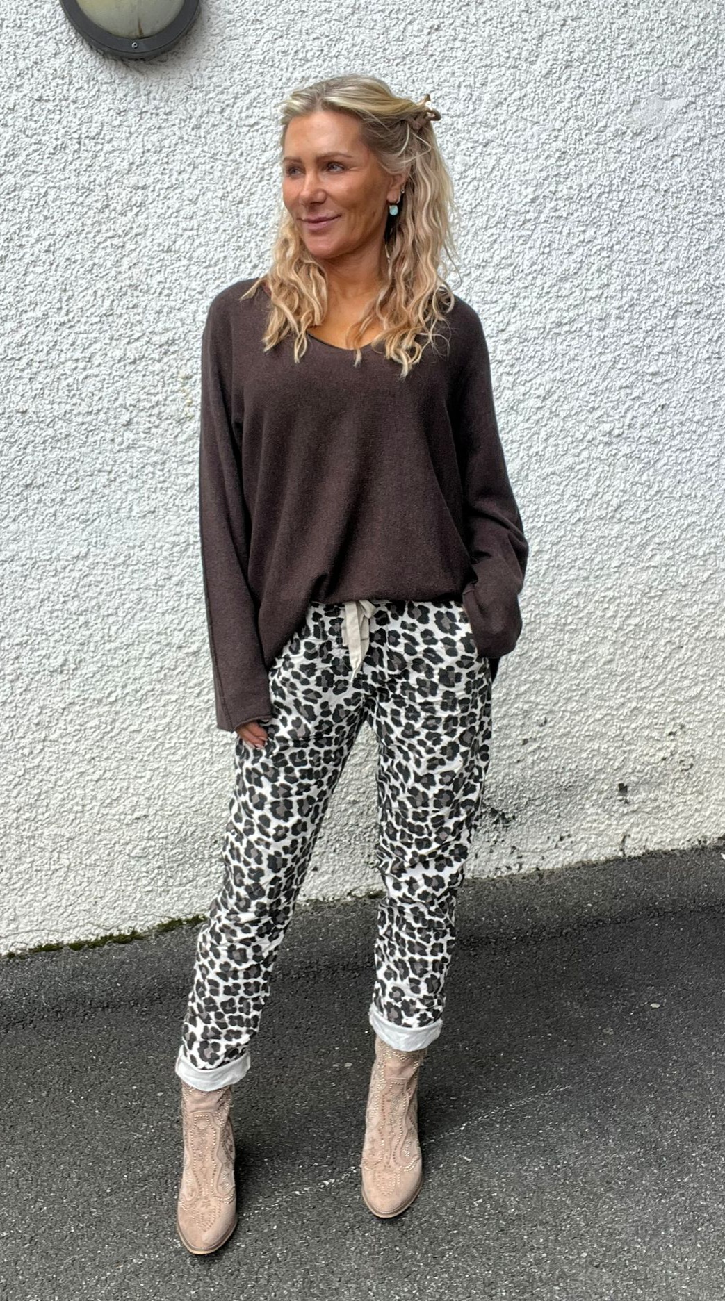 Blond kvinna står avslappnat mot en vit, grovputsad vägg och tittar åt sidan. Hon bär brun, lös tröja, leopardmönstrade byxor med uppvikta benslut och beige cowboyboots; ena handen i fickan.