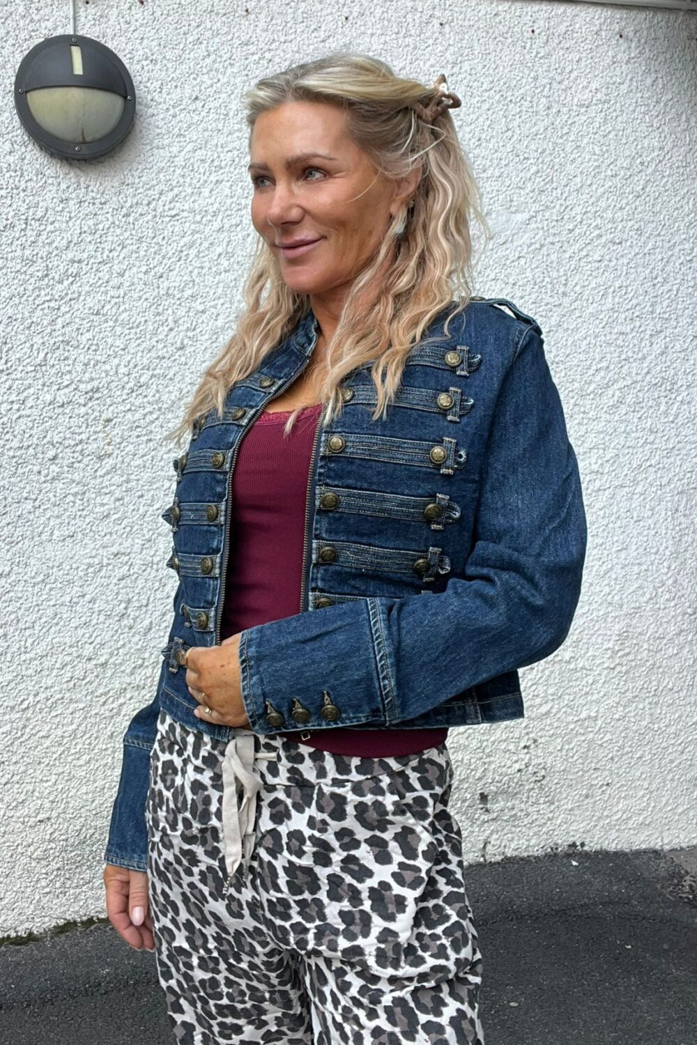 Blond kvinna står utomhus mot en vitputsad vägg och ler lätt medan hon tittar åt sidan. Hon bär kort blå jeansjacka med många knappar, vinröd topp och leopardmönstrade byxor; rund vägglampa syns bakom henne.