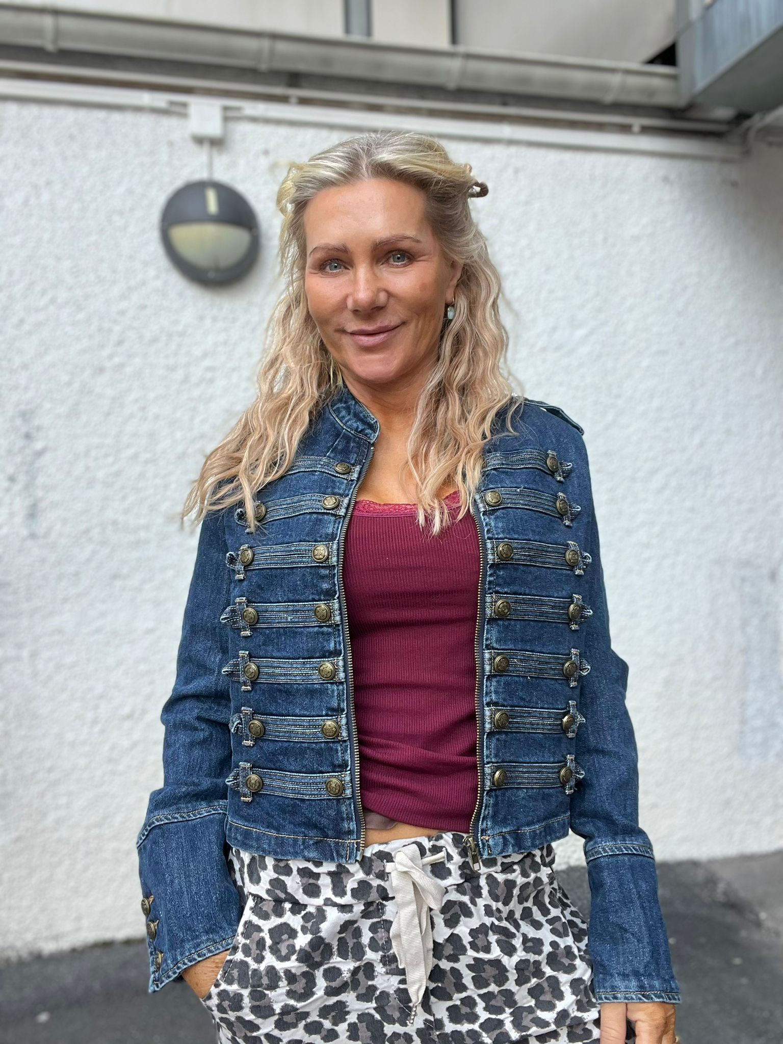 Leende kvinna med långt vågigt blont hår står och tittar mot kameran, klädd i blå jeansjacka med dekorativa knappar och vinröd topp. Hon står utomhus framför en ljus vägg med rund vägglampa.
