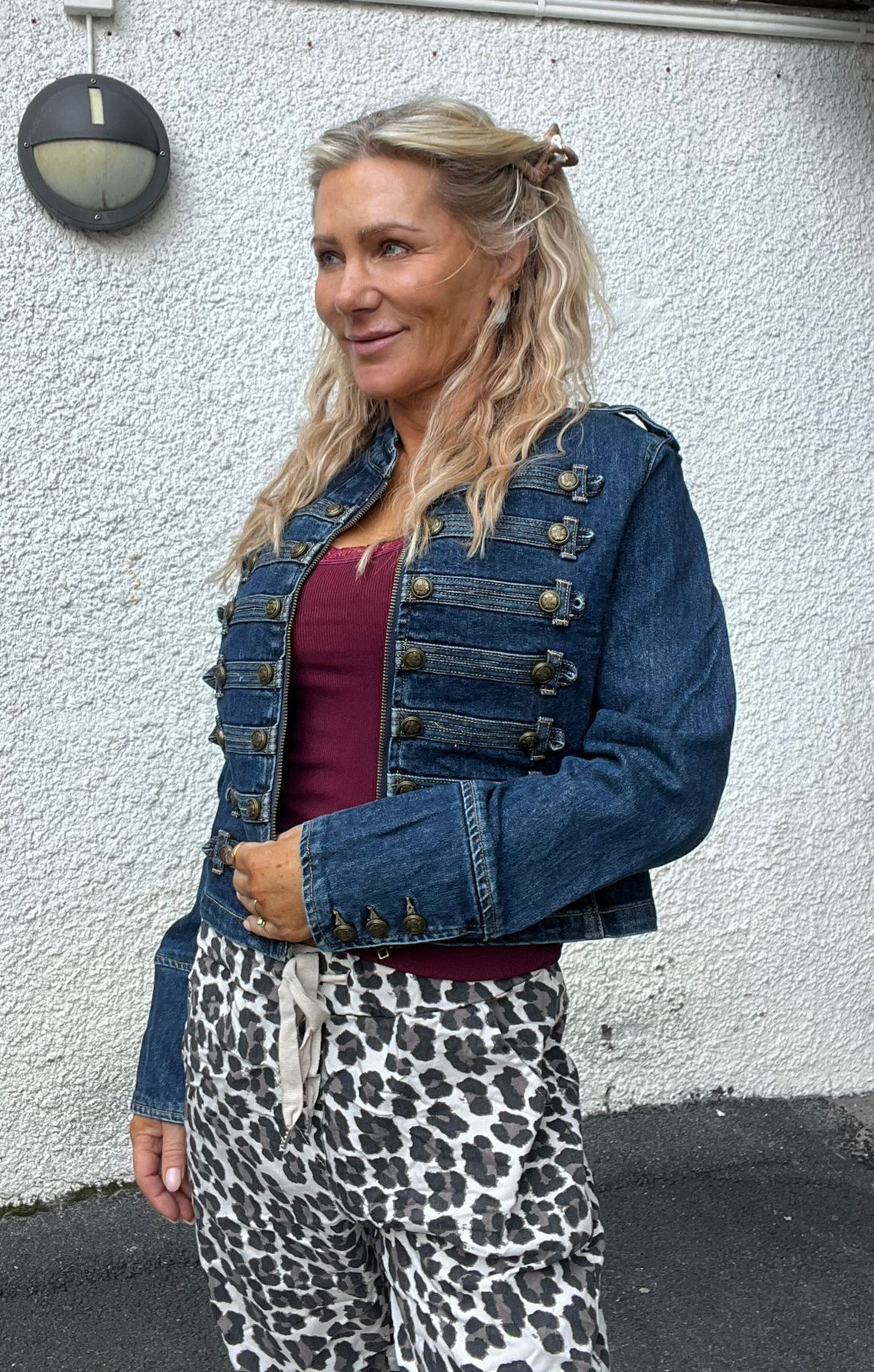Blond kvinna står utomhus mot en vitputsad vägg och ler lätt medan hon tittar åt sidan. Hon bär kort blå jeansjacka med många knappar, vinröd topp och leopardmönstrade byxor; rund vägglampa syns bakom henne.