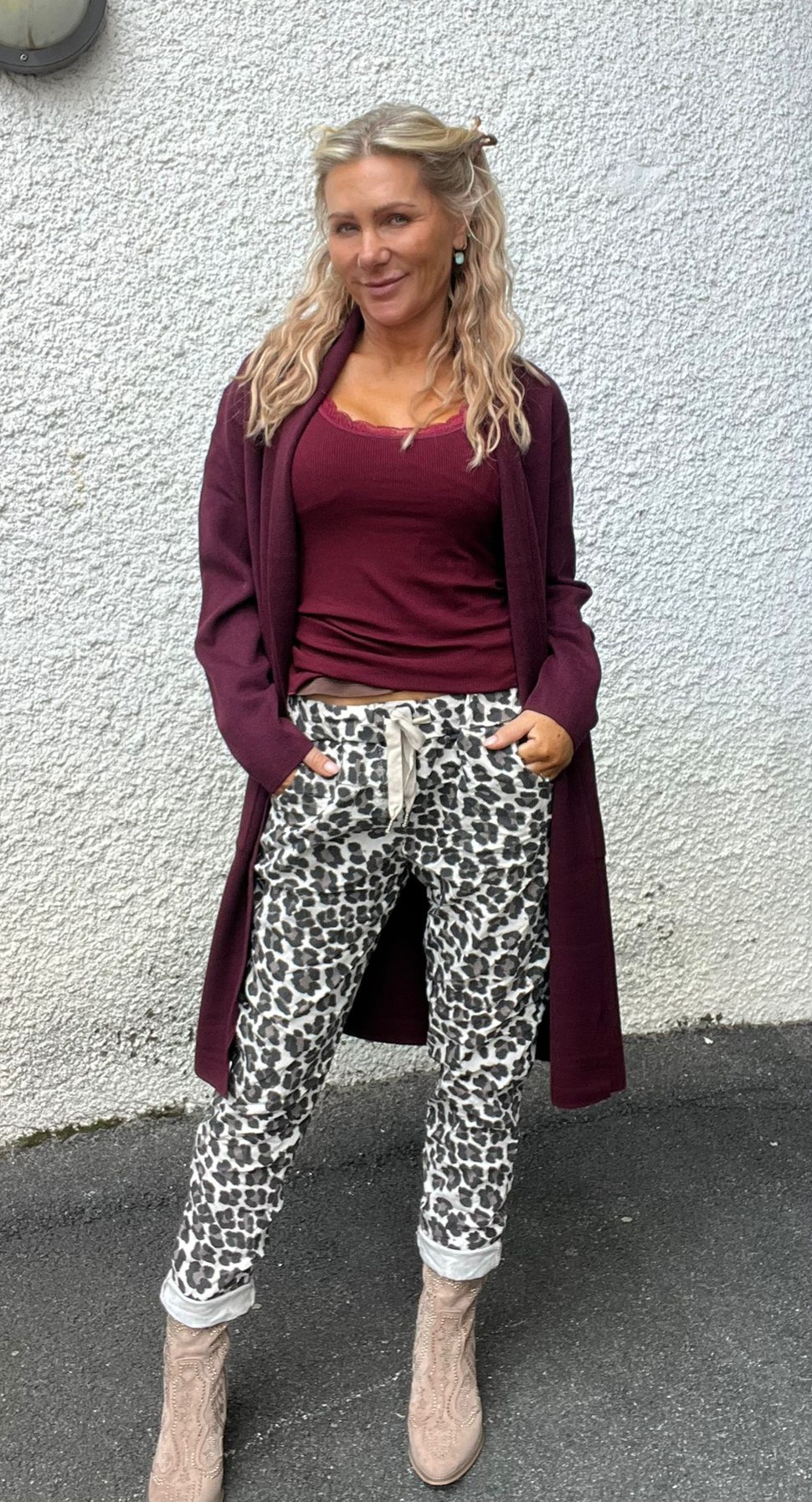 Kvinna med långt blont, vågigt hår står lutad mot en vit putsad vägg och ler mot kameran. Hon har vinröd topp och lång kofta, leopardmönstrade byxor och beige boots, med händerna i fickorna.