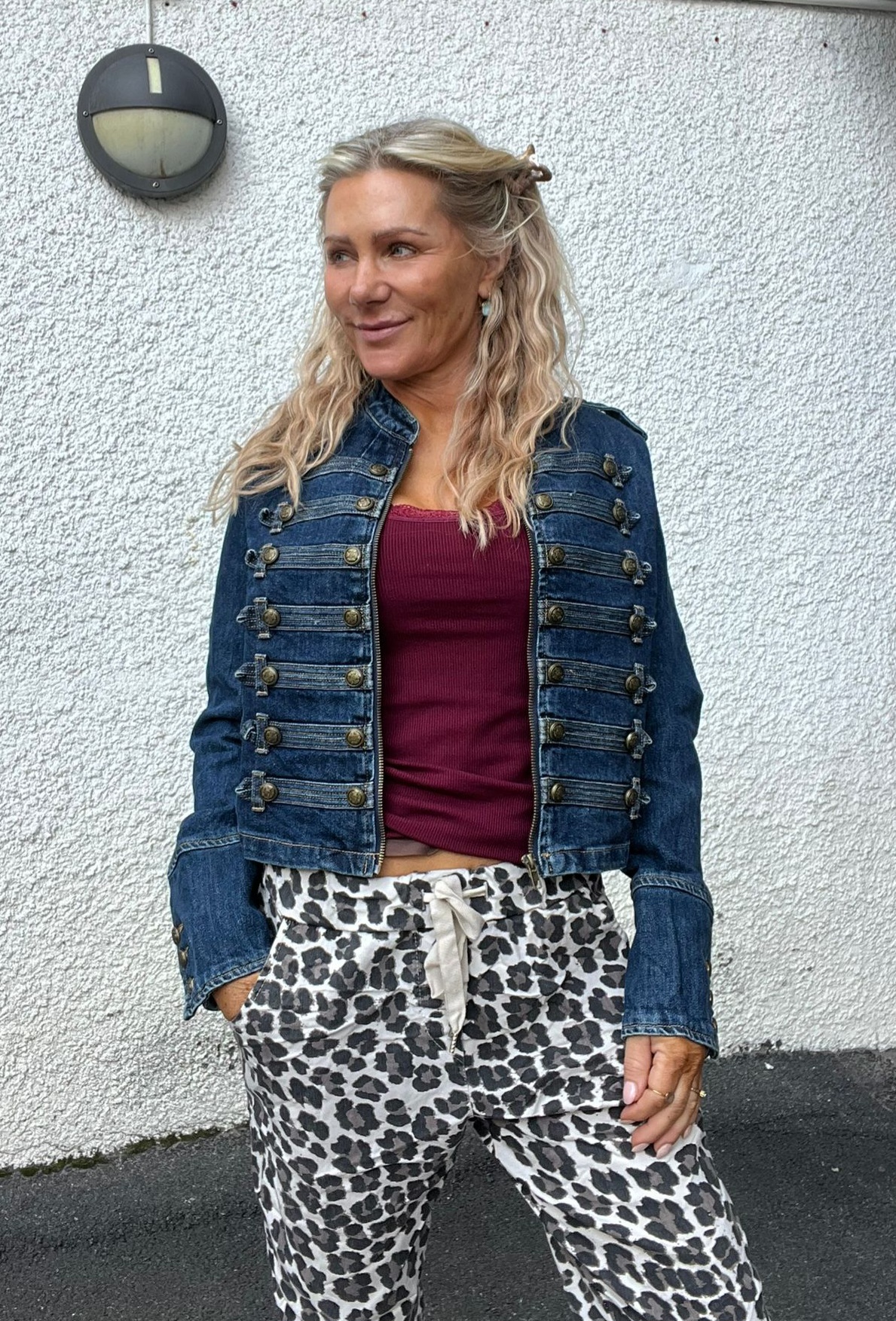 Kvinna med långt blont hår står avslappnat med handen i fickan och ler åt sidan. Hon bär jeansjacka med dekorativa knappar, vinröd topp och leopardmönstrade byxor, mot en vit putsad vägg med rund vägglampa.
