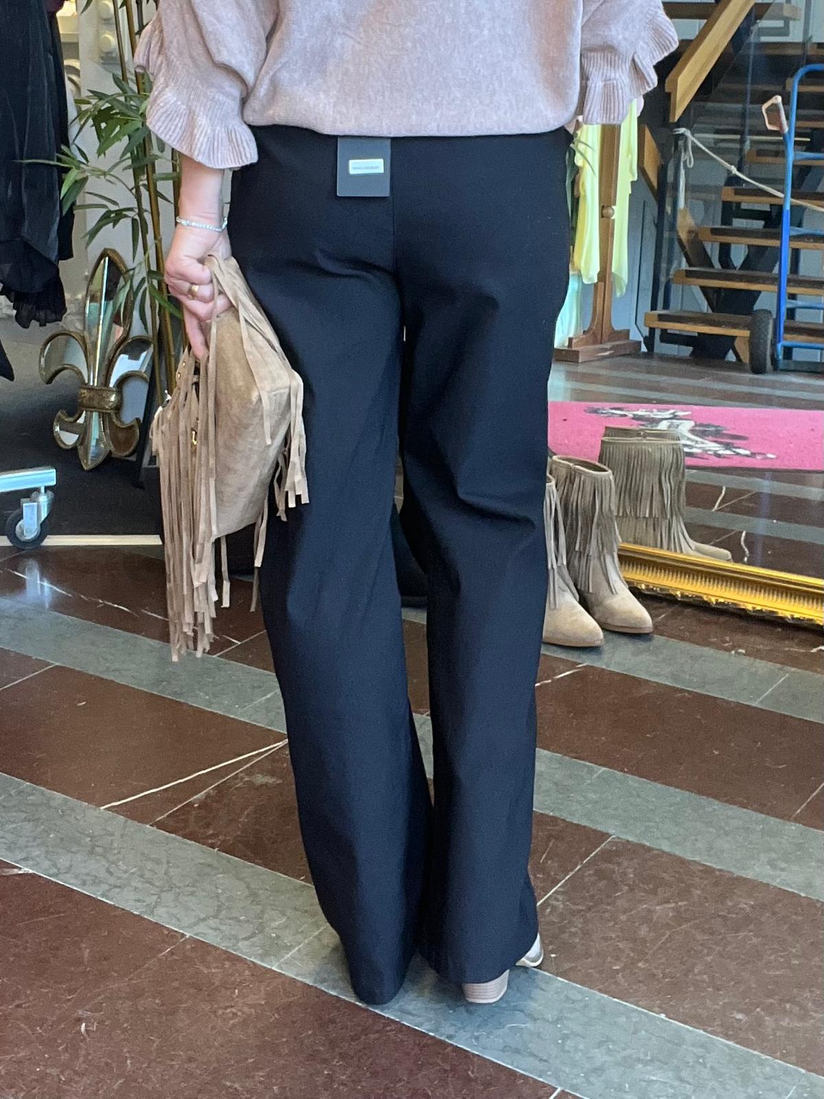 Person sedd bakifrån står i en butik och håller en beige fransväska i handen. Hen bär svart utsvängda byxor och en ljus blus med volangärmar; fransstövlar och klädställningar syns i bakgrunden.