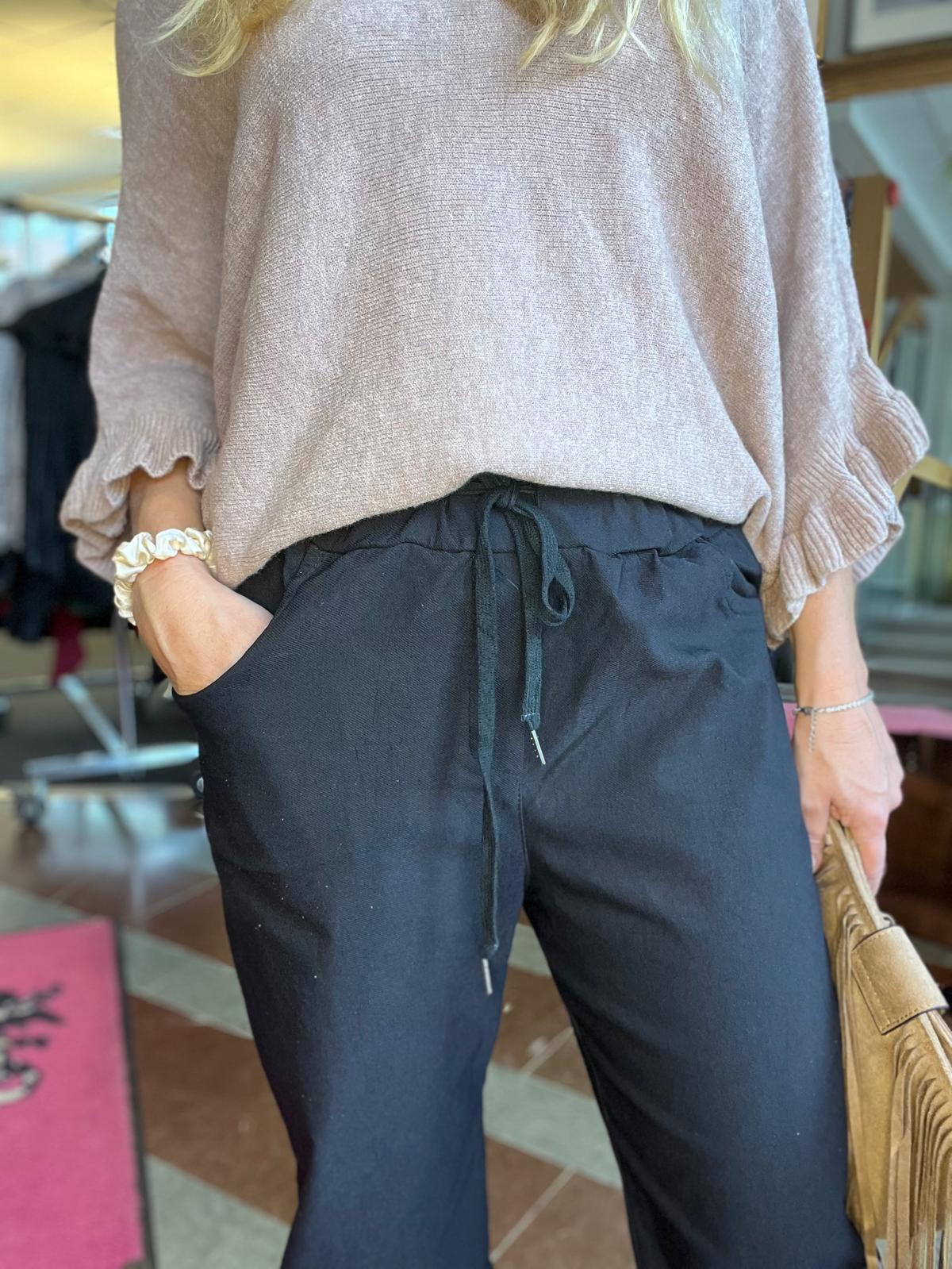 Person visar en vardaglig outfit, med handen i fickan. Hen bär en ljusbeige stickad tröja med volangärmar och svarta byxor med dragsko, samt håller en beige väska i en inomhusmiljö.