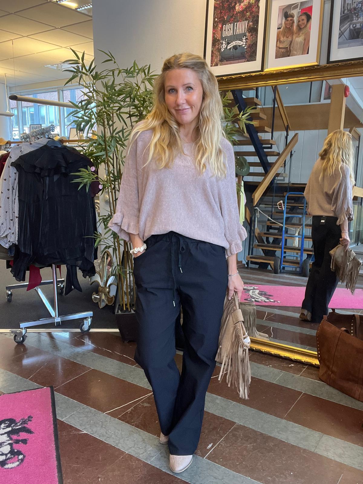 Kvinna med långt blont hår står och ler i en butiksmiljö, med händerna i fickorna och en beige fransväska i handen. Hon bär ljus stickad tröja och mörka byxor; spegel och klädställning syns bakom. Text: “GÅSA FANNY”.