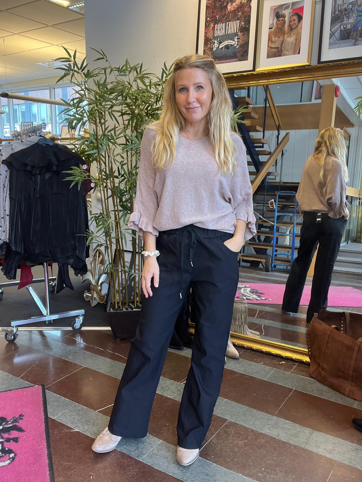 Blond kvinna står och poserar med ena handen i fickan, klädd i ljusrosa tröja, svarta byxor och ljusa sneakers. Hon är i en klädbutik vid en stor spegel, klädställning och växt; skylt text: “GÅSA FANNY”.