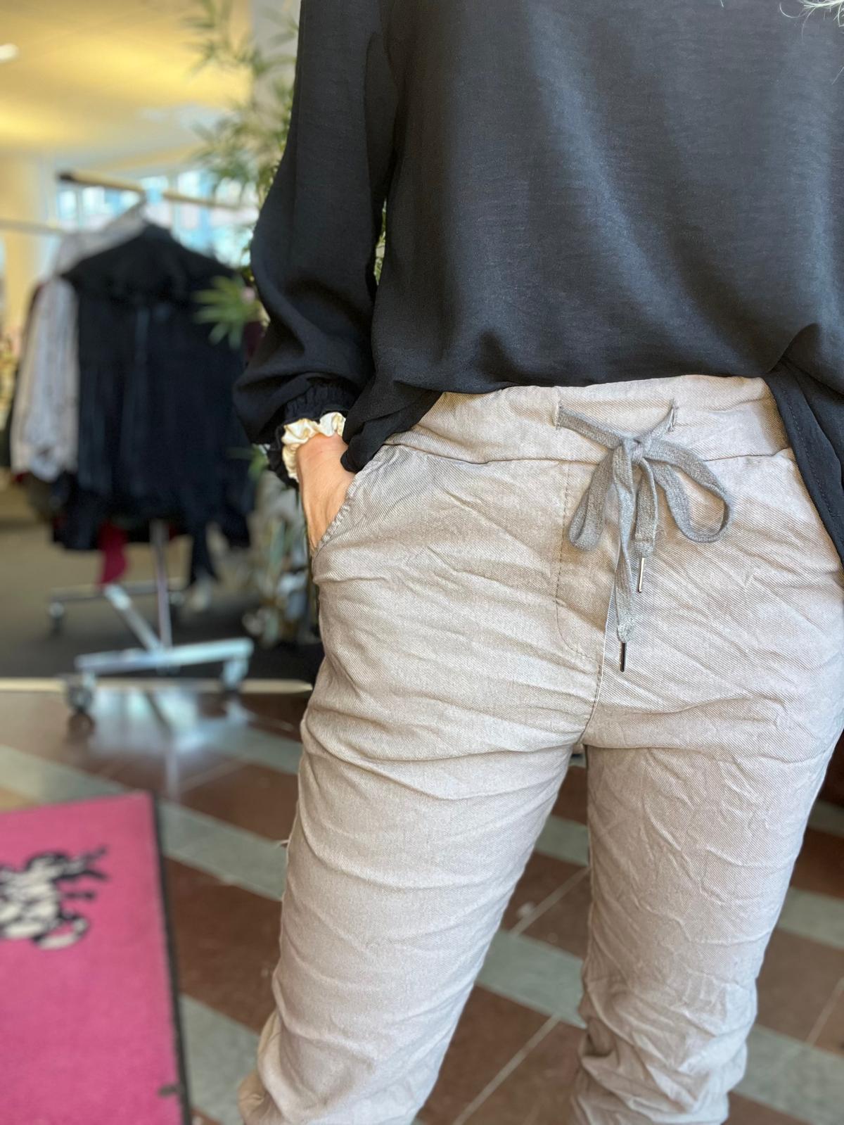 Person står med handen i fickan och visar ljusbeige byxor med knytband i midjan och svart blus. Bakgrunden är suddig men liknar en klädbutik med klädställning och växt.
