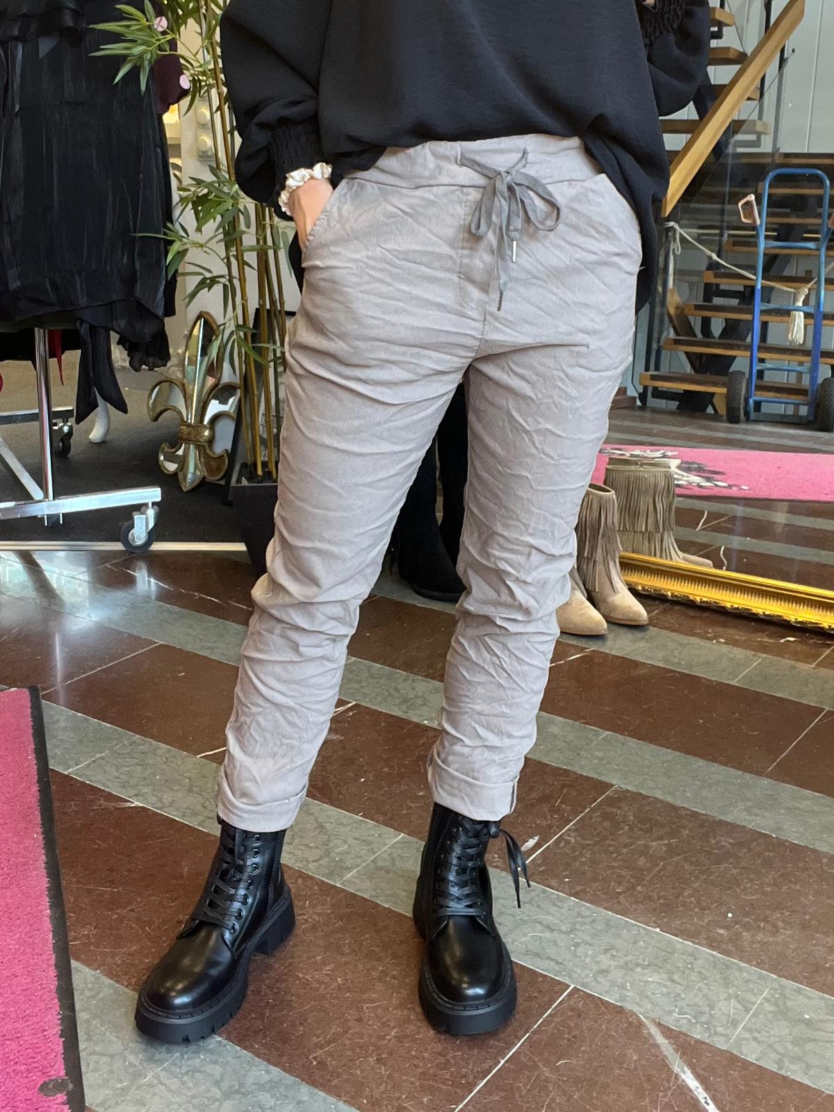 Person står avslappnat med handen i fickan och visar ljusgrå byxor med dragsko i midjan och svarta snörkängor. Miljö i en butik eller studio med klädställning, växt och trappa i bakgrunden.