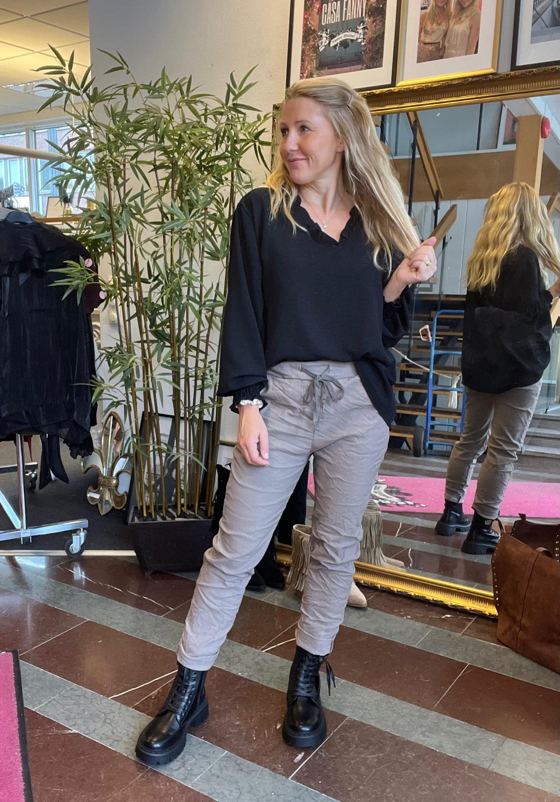 Blond kvinna poserar inomhus och håller upp en hårslinga medan hon tittar åt sidan. Hon bär svart blus, grå byxor och svarta kängor, står vid en stor spegel med bambuplanta, klädställning och väskor. Text: “CASA FANNY”.