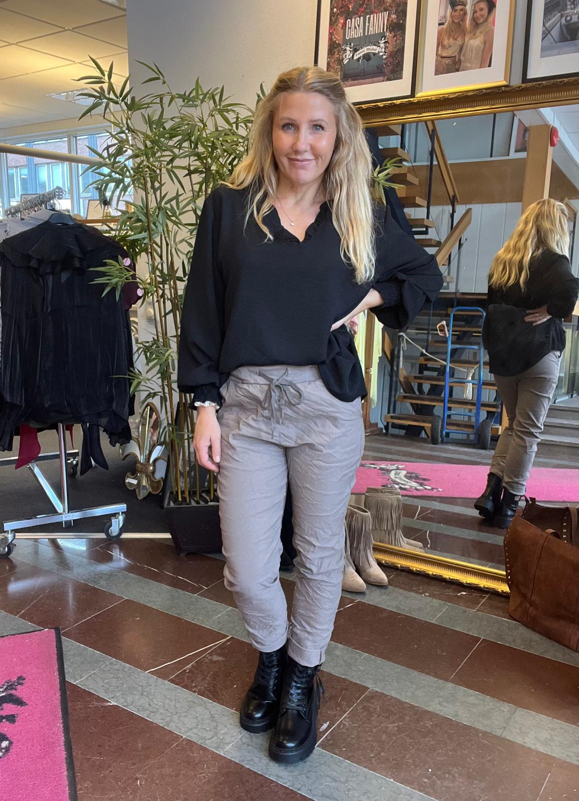 Blond kvinna står och poserar med handen på höften i en butikslik miljö, klädd i svart blus, grå byxor och svarta kängor. Bakom syns spegel, klädställning, trappa samt affischtexten “CASA FANNY”.