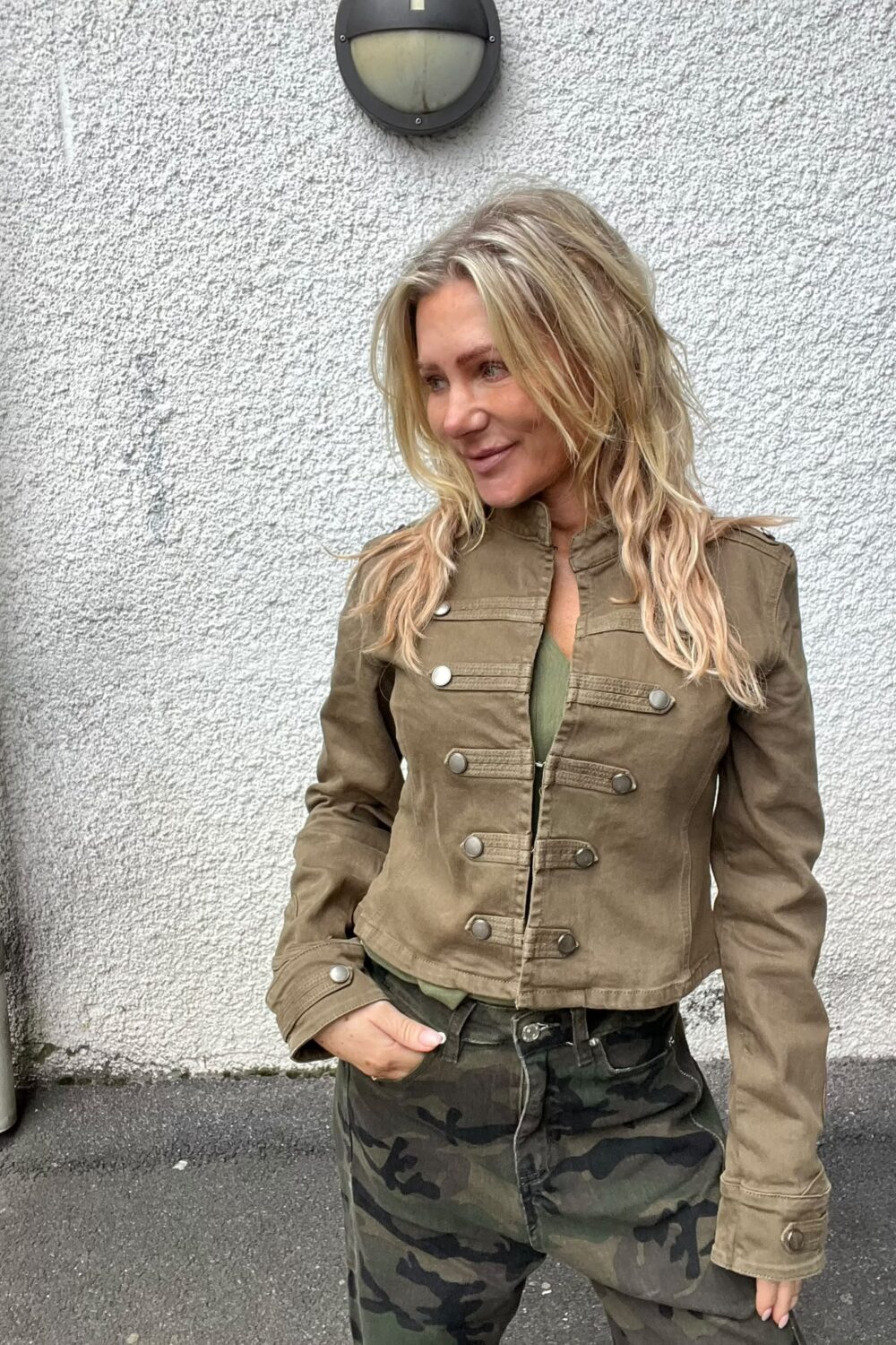 Blond kvinna står lutad mot en vit, grovputsad vägg och ler medan hon tittar åt sidan. Hon har en brun jacka och camouflagemönstrade byxor, med en hand i fickan och en vägglampa ovanför.