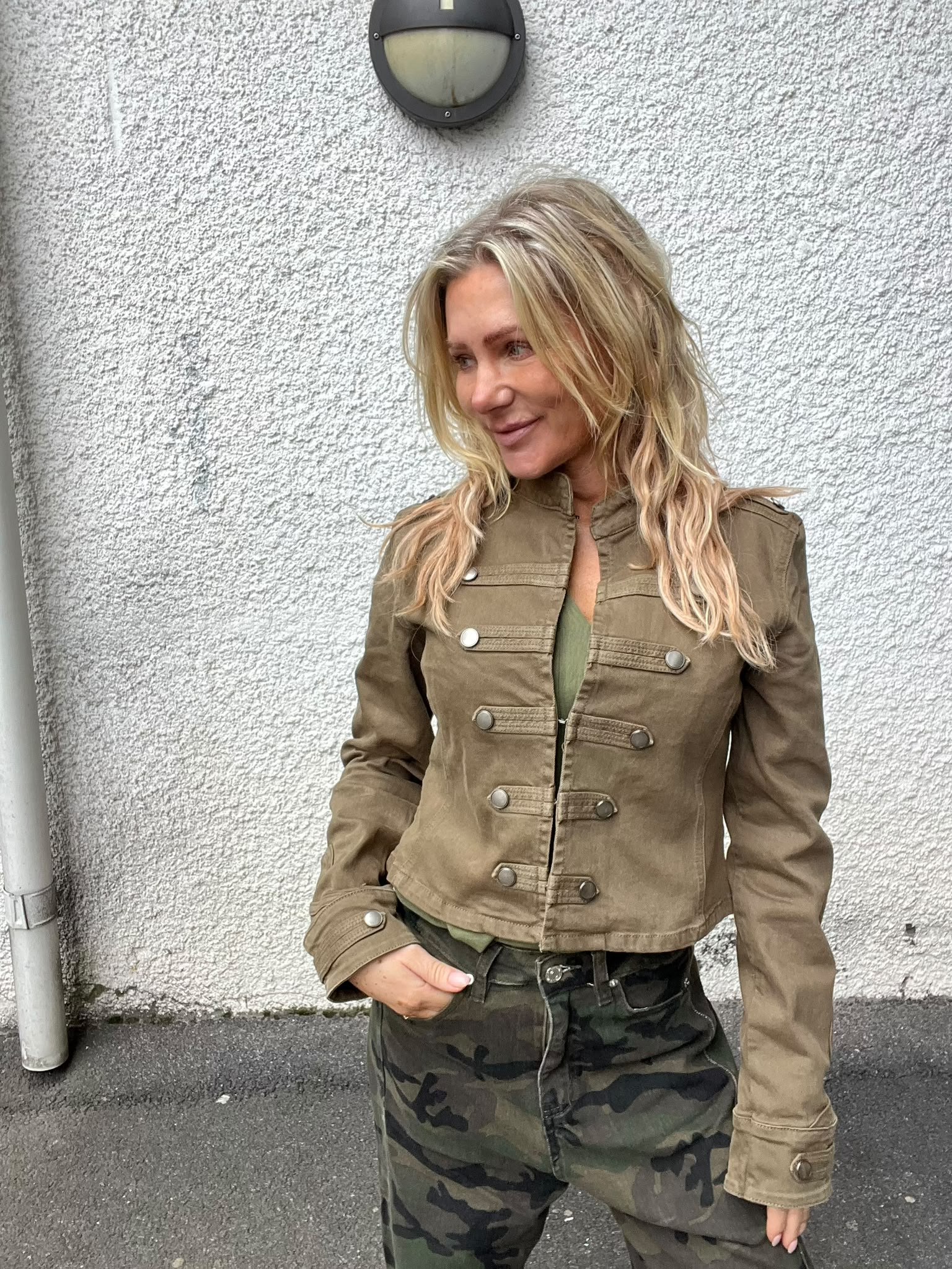 Blond kvinna står lutad mot en vit, grovputsad vägg och ler medan hon tittar åt sidan. Hon har en brun jacka och camouflagemönstrade byxor, med en hand i fickan och en vägglampa ovanför.